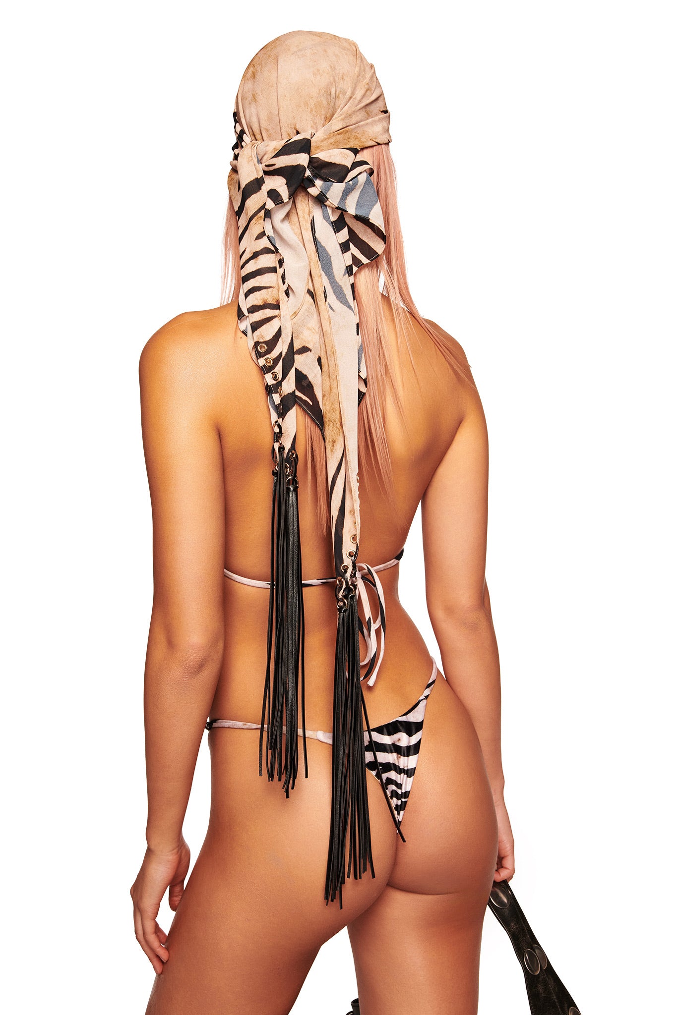 A person with long light pink hair, seen from behind, wears a zebra-print bikini and the I.AM.GIA ALLUNA MULTI-WEAR TASSEL SCARF - WILD SIDE adorned with vegan suede tassels, holding an object partially visible on the right.