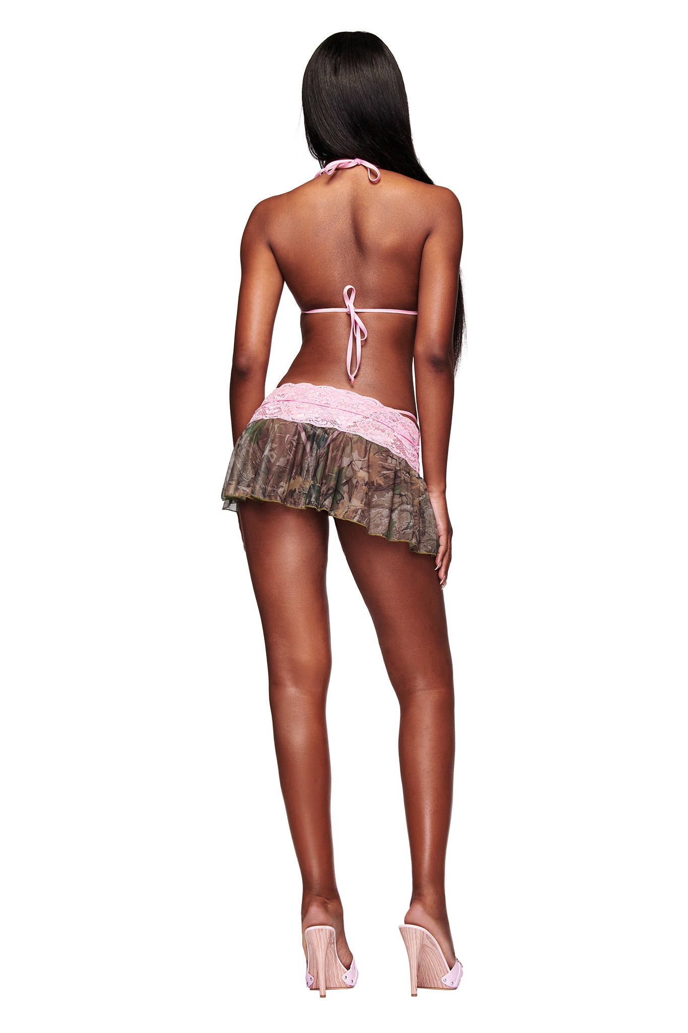 A woman with long dark hair faces away from the camera, wearing the I.AM.GIA SAKINA BIKINI TOP - BARK AND BITE CAMO and a short camo print mini skirt with high heels against a white background.