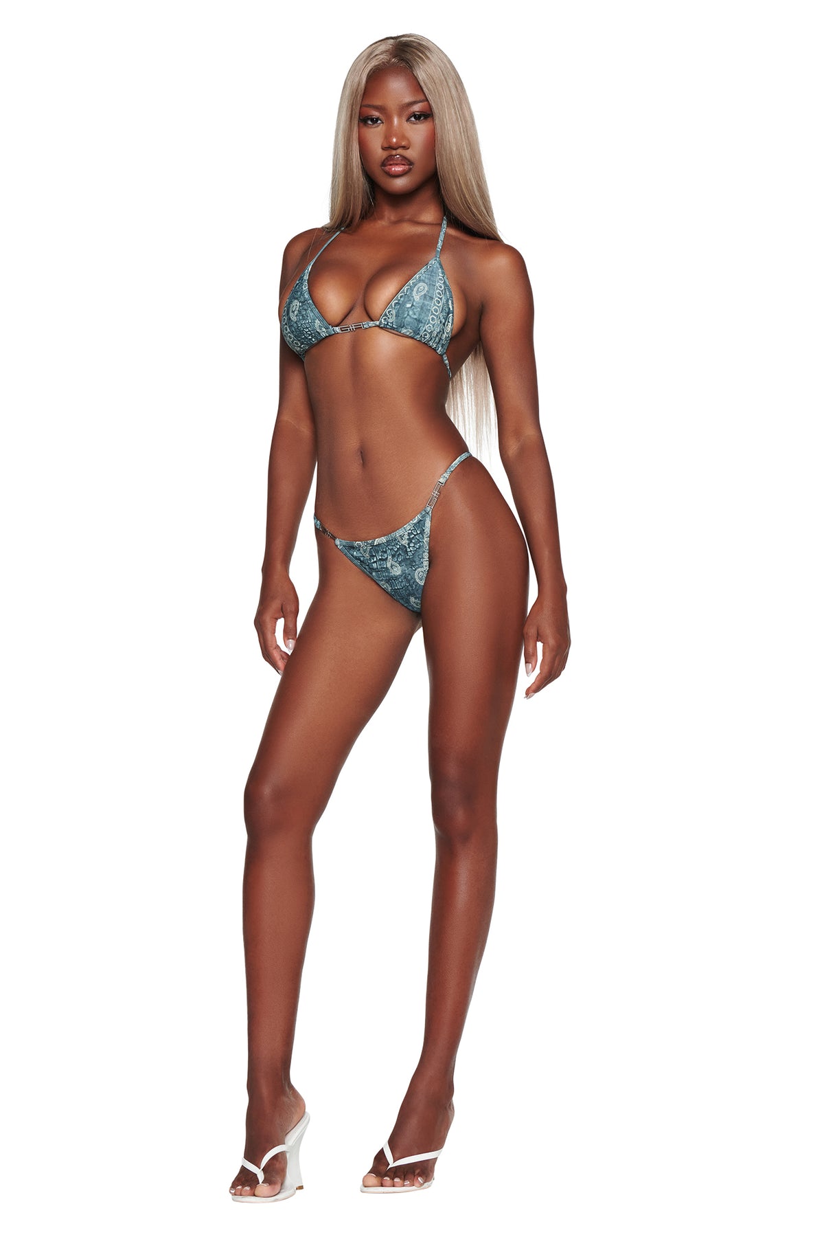A woman with long blonde hair stands against a white background wearing the I.AM.GIA T-REX AVA BIKINI BOTTOM in blue bandana print paired with a matching bikini top and clear-heeled sandals.