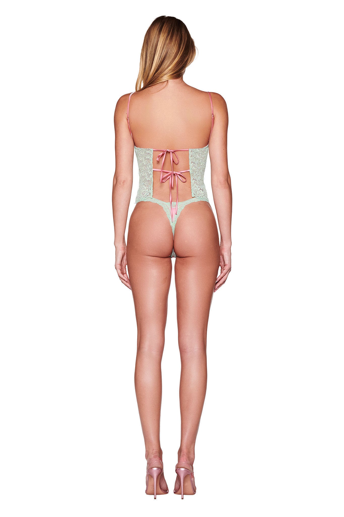 A person with long hair stands with their back to the camera, highlighting the LOLO 2 PIECE SET - PINK from I.AM.GIA—a pink and green lace bodysuit adorned with a ribbon tie, enhanced by sheer lace details and paired with pink high heels. The plain white background emphasizes the elegance of the ensemble.