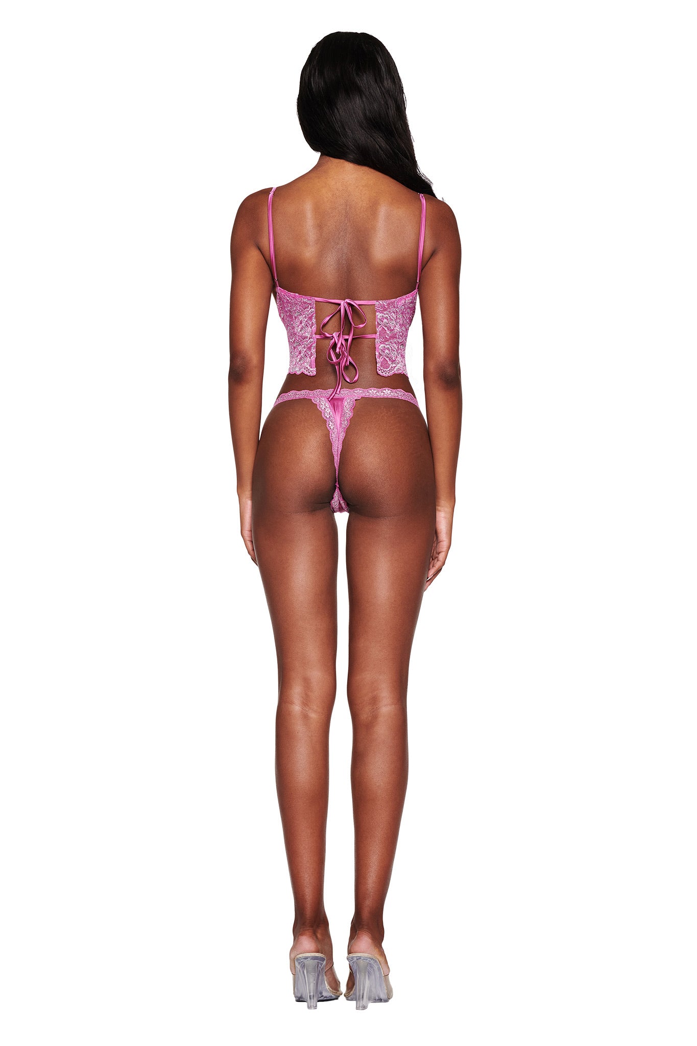 A person is seen from the back wearing the LOLO METALLIC SET - MAGENTA by I.AM.GIA, featuring a magenta metallic lace corset with adjustable straps and ribbon details, paired with a matching low rise thong, standing on a white background.