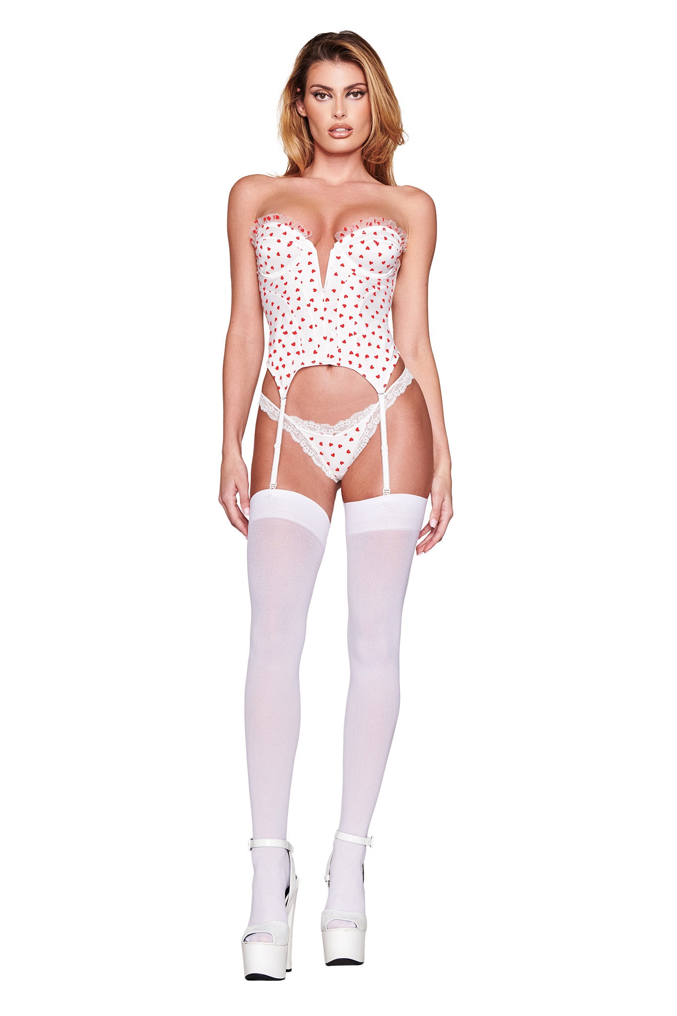 A person models the PAMELA SET - WHITE : HEARTBEAT by I.AM.GIA, featuring a vintage-inspired pink strapless corset with red heart patterns over heart flocked tulle, a matching thong, white thigh-high stockings, and white platform heels against a plain white background.