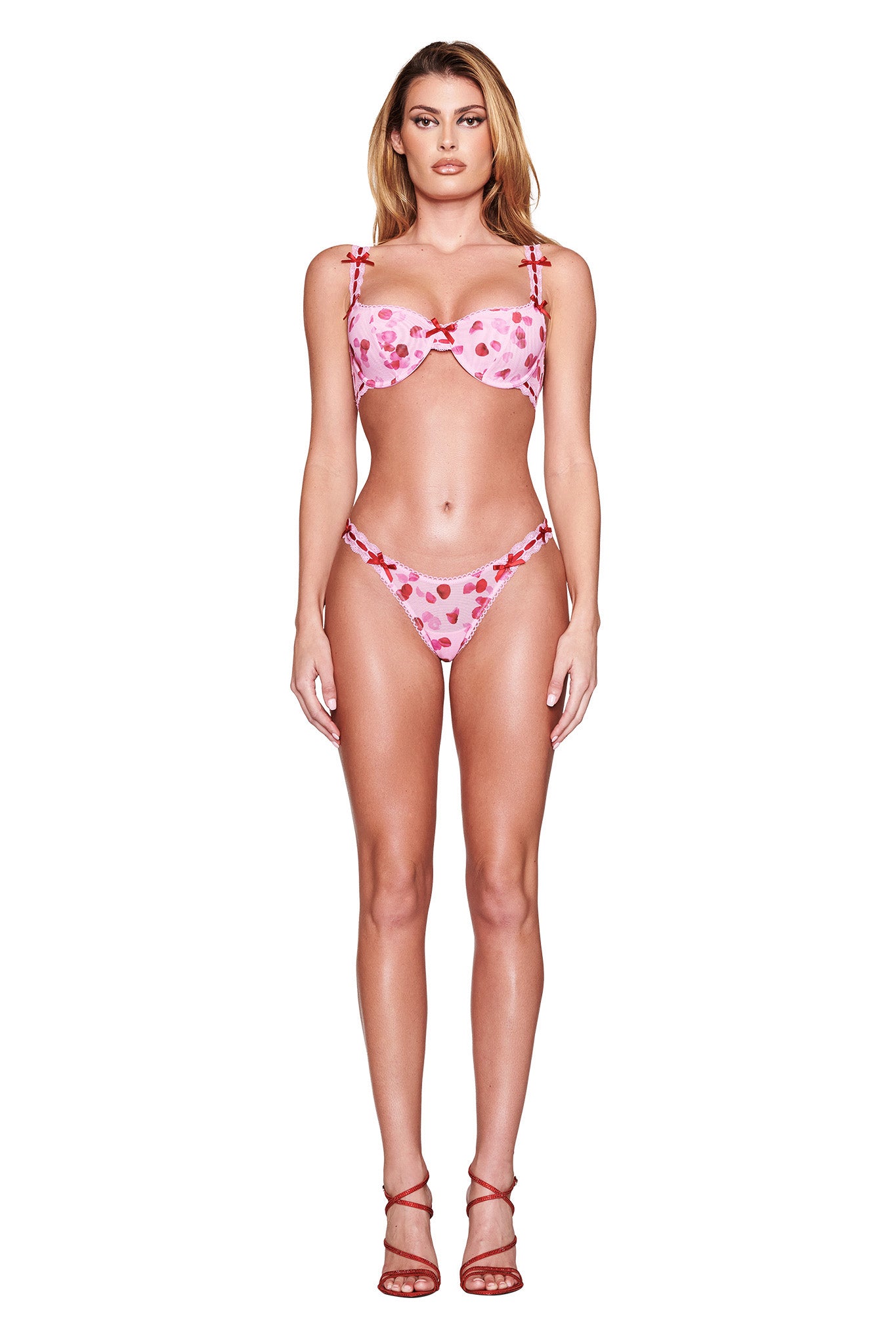 A woman with long, wavy hair faces forward in the pink GABRIELLA 2 PIECE SET - PRETTY PETAL by I.AM.GIA, featuring heart patterns and adjustable straps. She's wearing red sandals against a plain white background that beautifully highlights the swimwear.