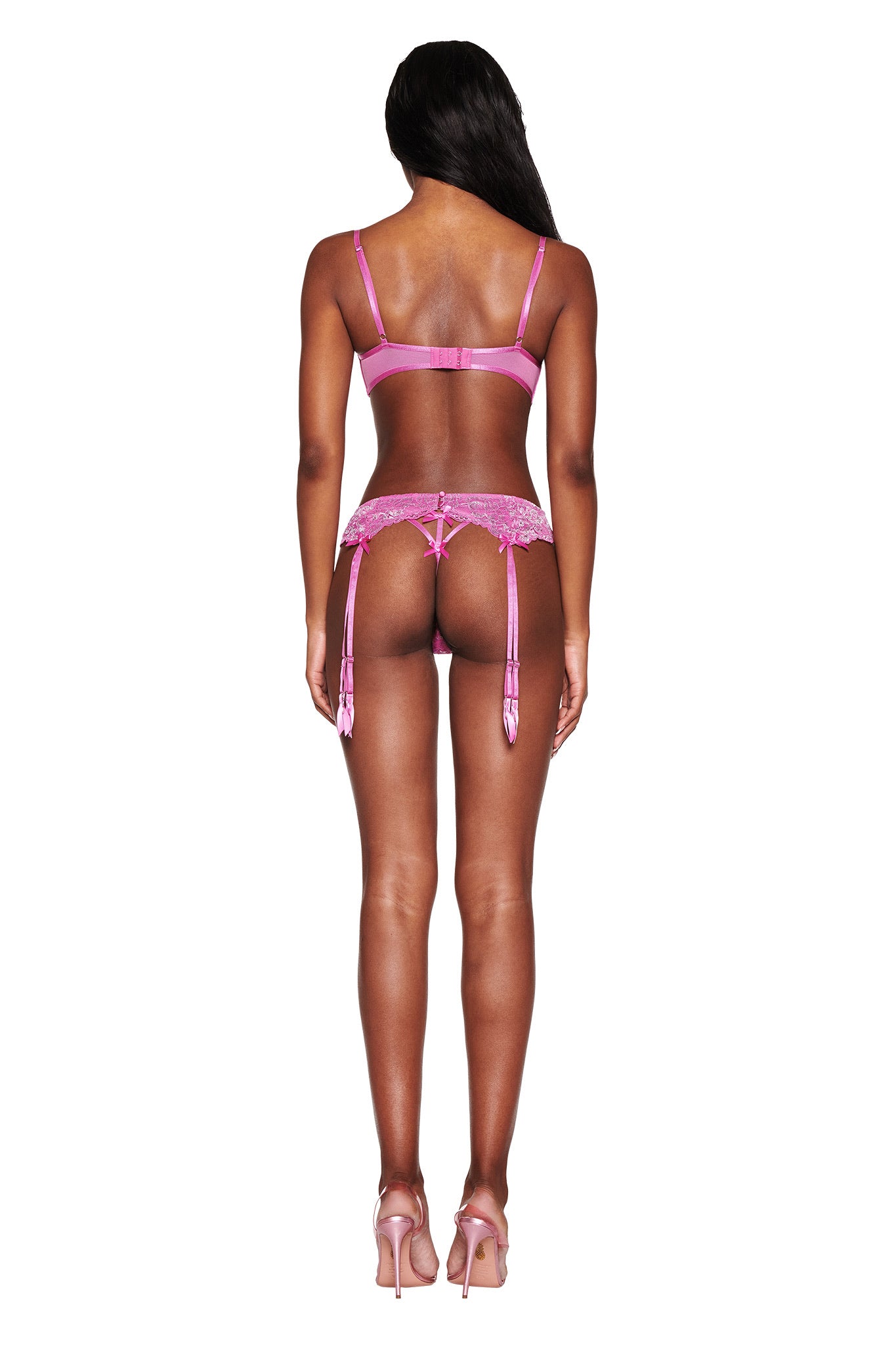 A person in the Y2K-inspired RAINE 3 PIECE METALLIC SET - PINK from I.AM.GIA, featuring pink lingerie with garter straps, stands facing away from the camera. Long black hair and pink high-heeled shoes complement the look against a plain white background.