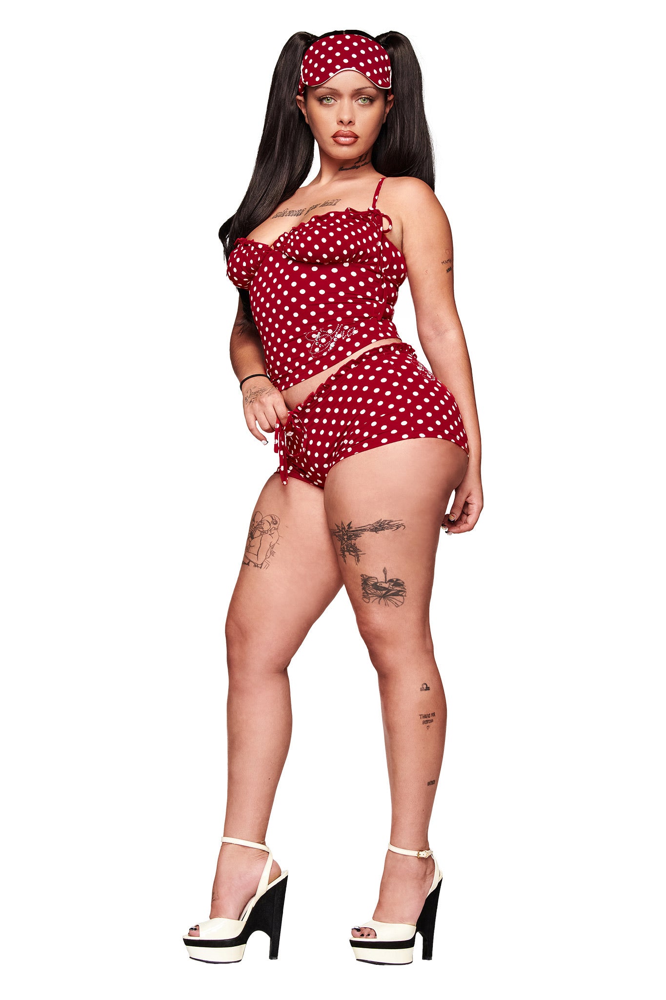 A woman wears the I.AM.GIA MELANIE SHORT - RED POLKA DOT set with micro mini shorts, an eye mask, white platform heels, long dark pigtails, and shows tattoos on her legs and arm as she poses confidently.