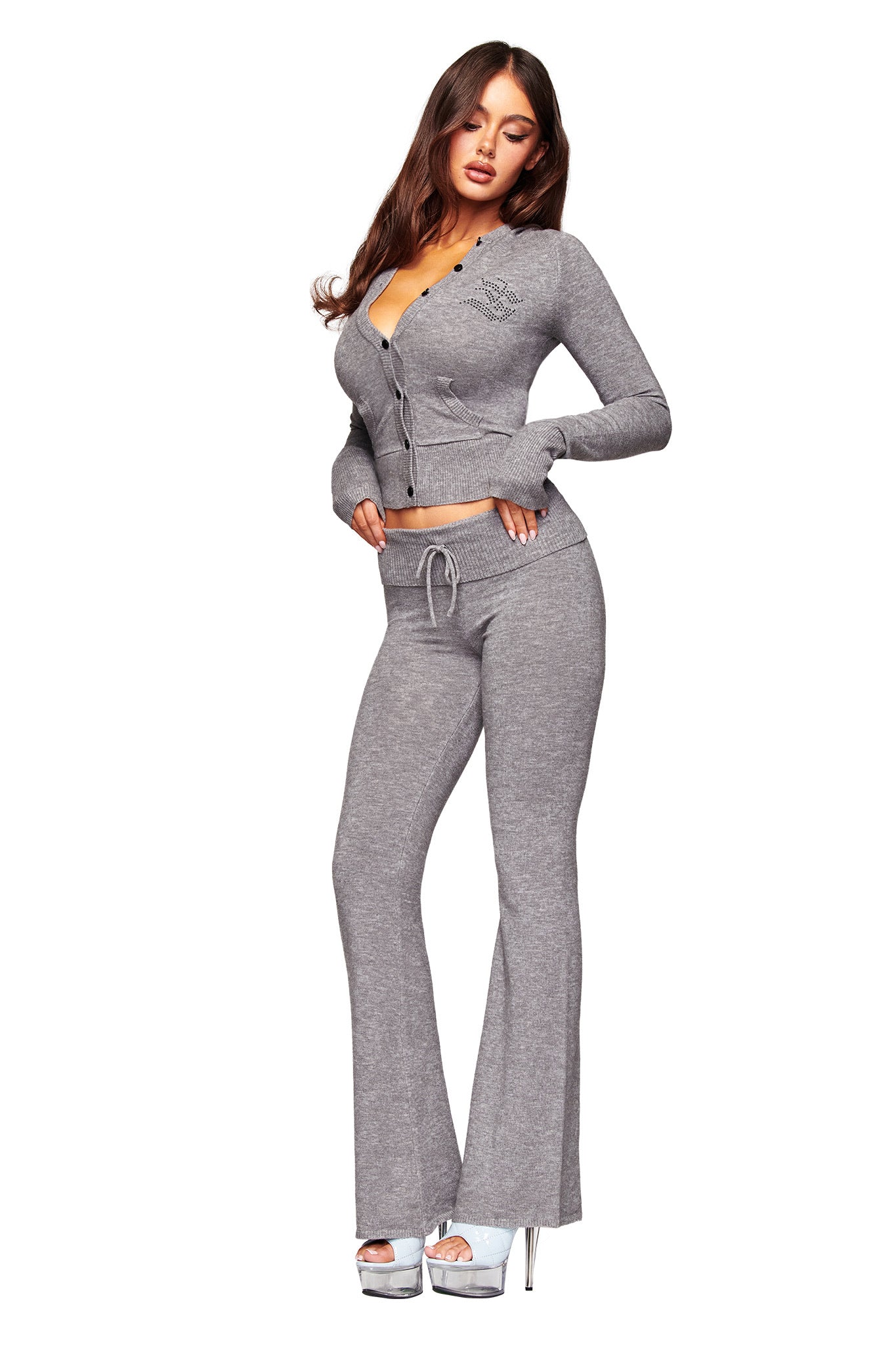 A woman poses in the I.AM.GIA JESSA PANT - GREY, paired with a light gray knit cardigan and silver platform heels. Her long, wavy brown hair flows elegantly against a white background.