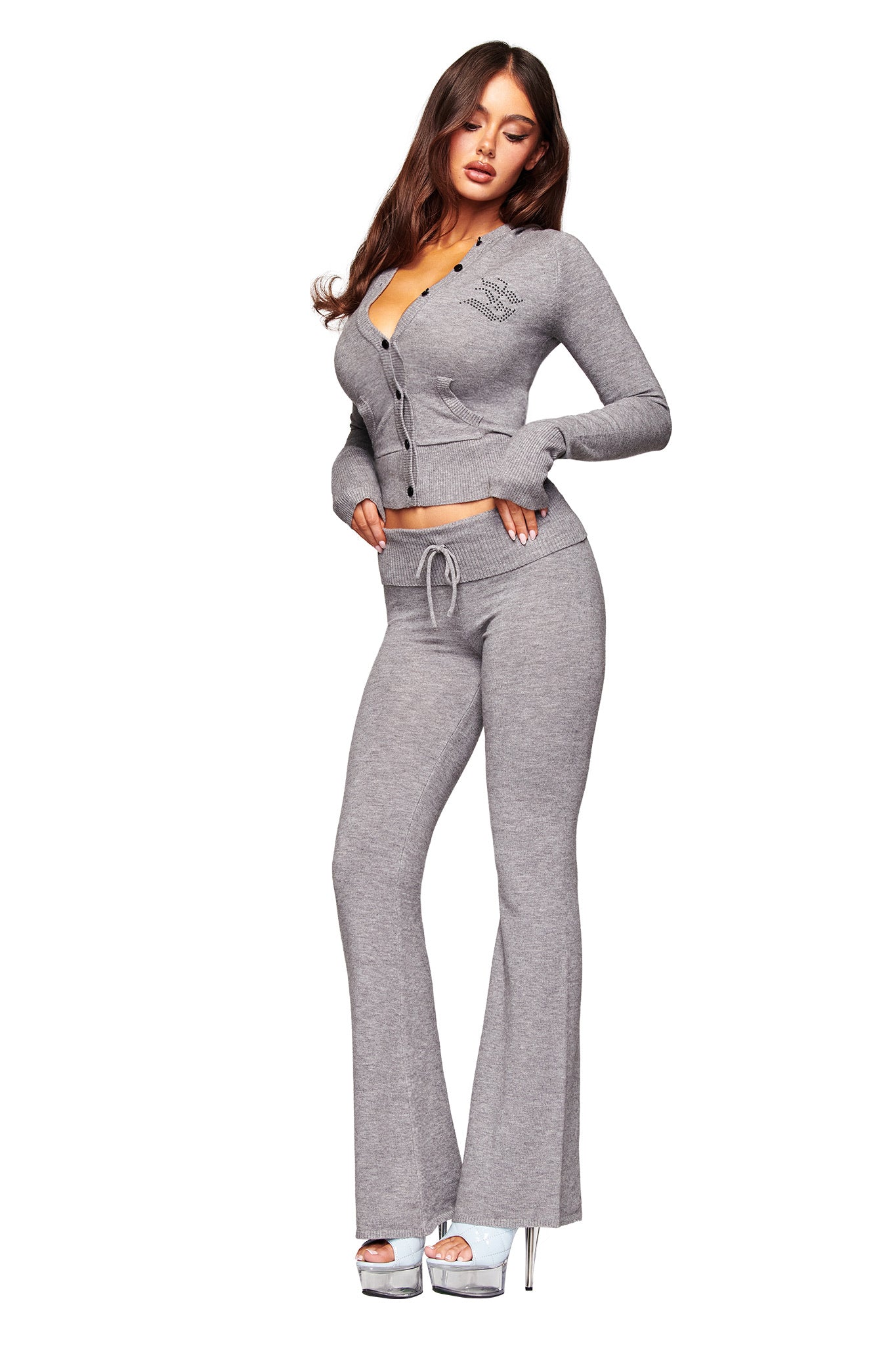 A woman poses in the I.AM.GIA JESSA HOODIE in grey, a fitted knit two-piece with a buttoned cardigan and drawstring pants, styled with silver platform heels against a white background.