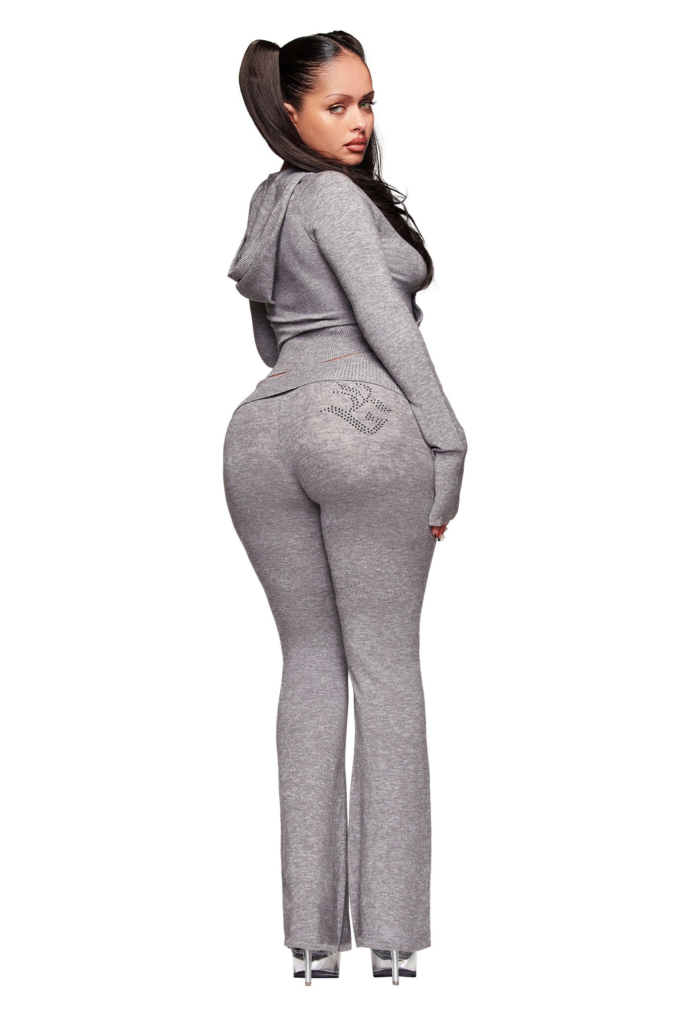 A woman wearing the I.AM.GIA JESSA PANT in grey, paired with a fitted, light gray hooded sweater with rhinestone details, stands facing away and looks over her shoulder. She has long dark hair in a ponytail and wears high-heeled shoes.
