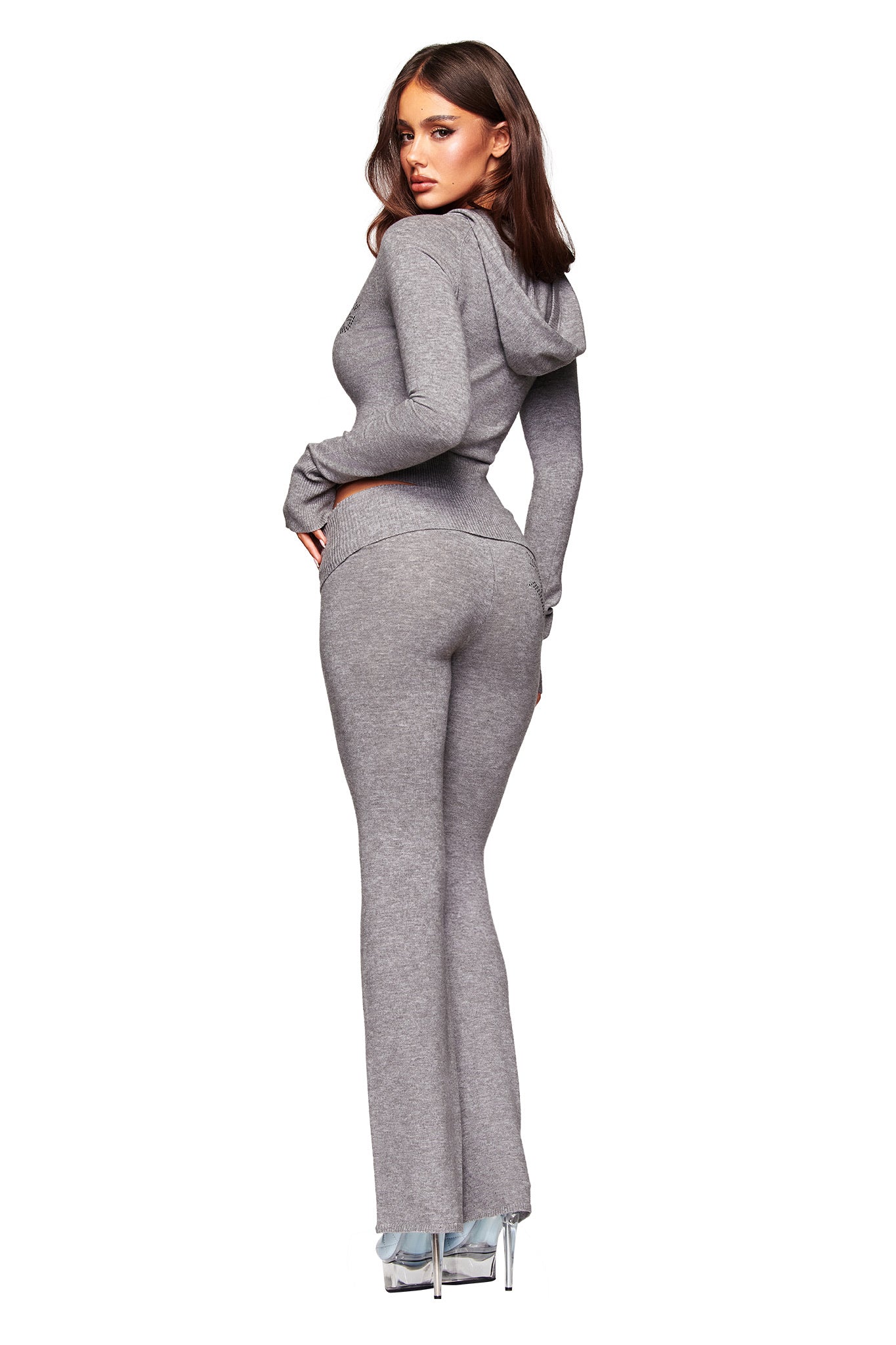 A woman models the I.AM.GIA JESSA PANT - GREY, a charcoal lounge pant with a mid-rise waist, paired with a fitted gray hoodie and clear high heels. She stands back to the camera, glancing over her shoulder against a white backdrop.