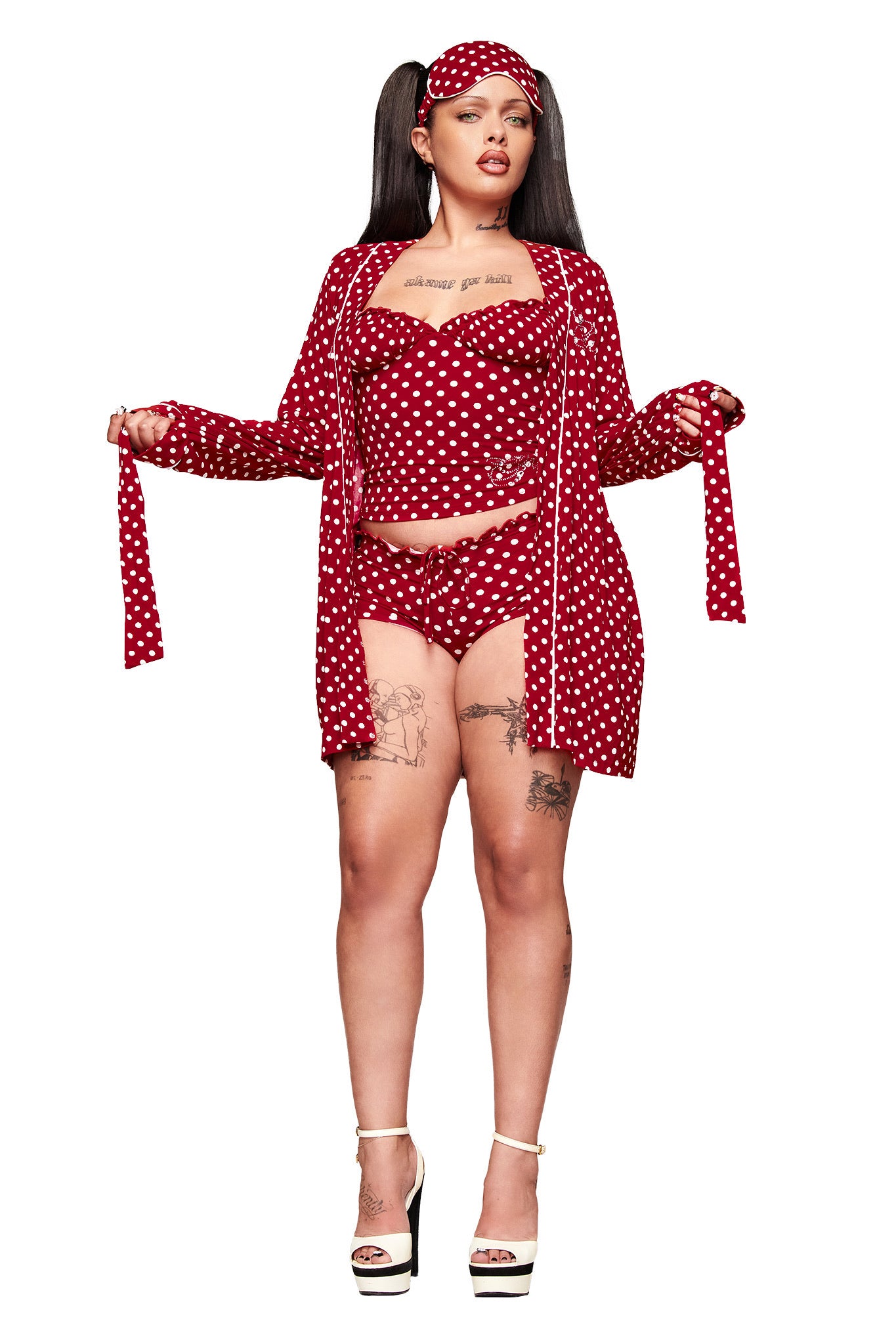 A woman wears the I.AM.GIA MELANIE SLEEPWEAR - RED POLKA DOT set with a matching robe and eye mask. She stands confidently in white platform heels, showing off leg tattoos and pigtails against a white background.