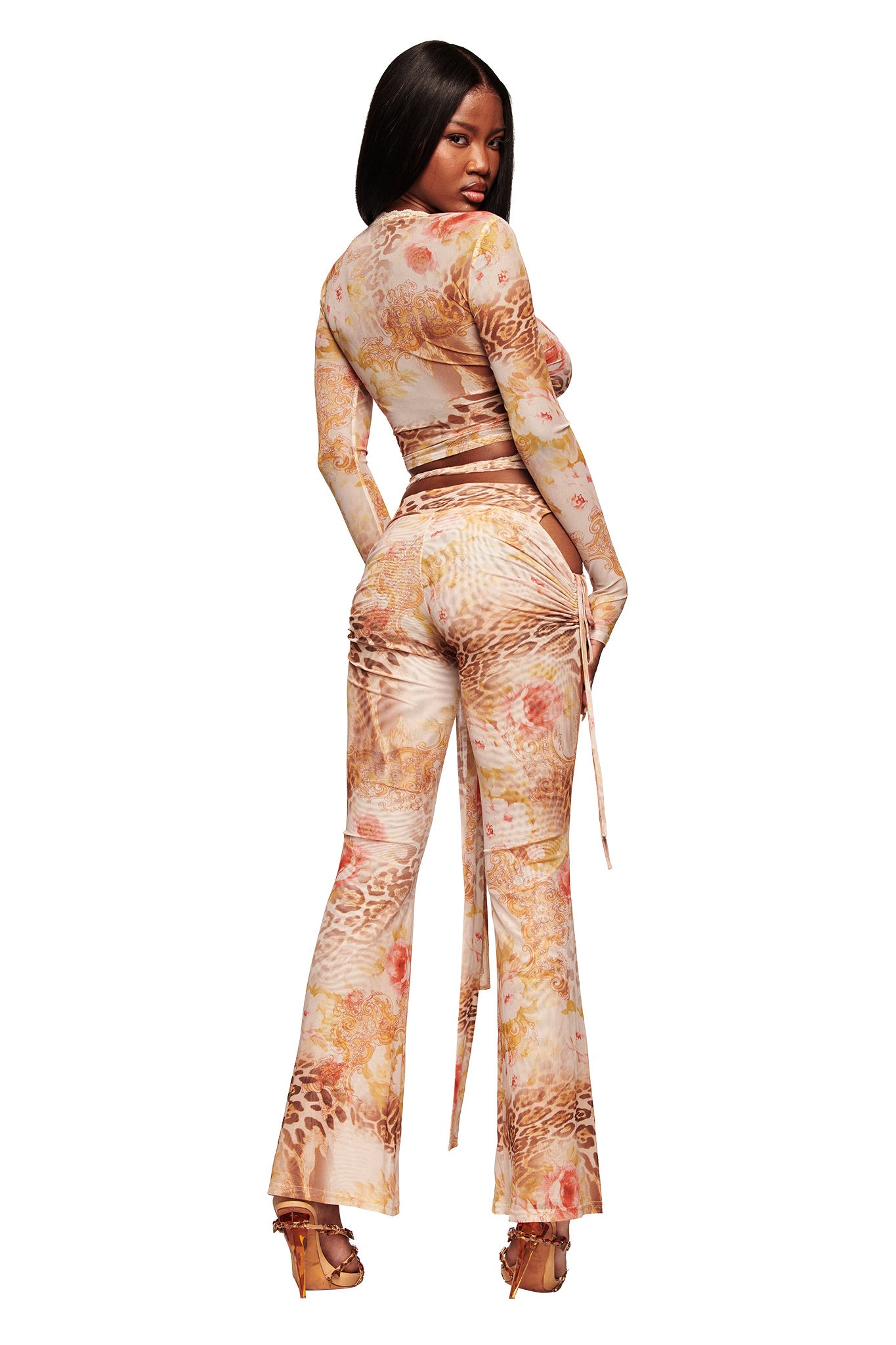 A woman with long, straight black hair poses in the I.AM.GIA ERISKA TOP - DESERT ROSE, a fitted mesh wrap top with a beige and brown floral-animal print, paired with flared pants and high-heeled sandals as she looks back over her shoulder.