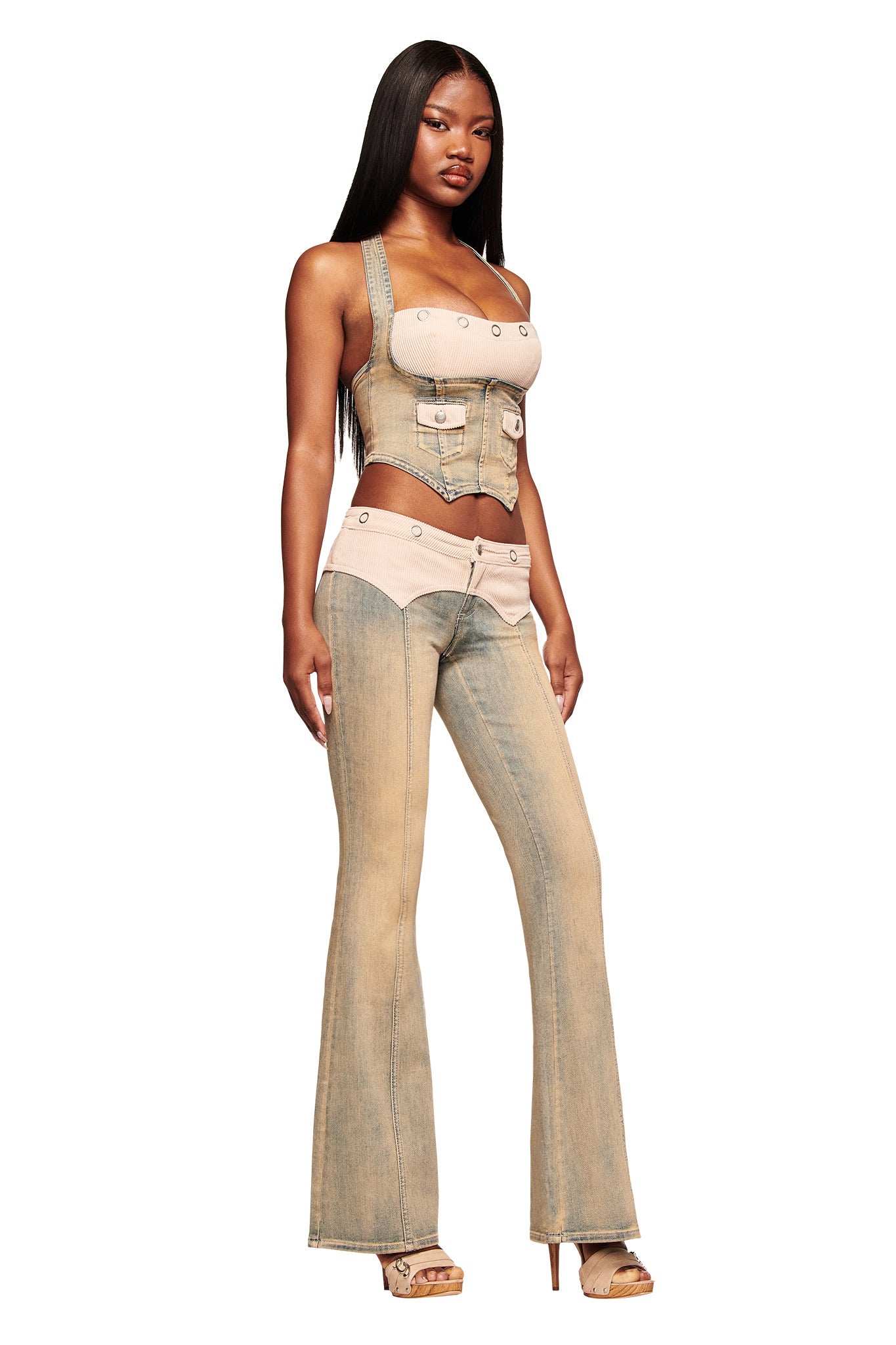 A woman stands confidently in the I.AM.GIA TOMMIE JEAN - ANTIQUE DENIM, featuring flared legs, beige accents, and a mid rise waist, paired with a fitted sleeveless corset top and high-heeled sandals. Her long straight hair completes the look.
