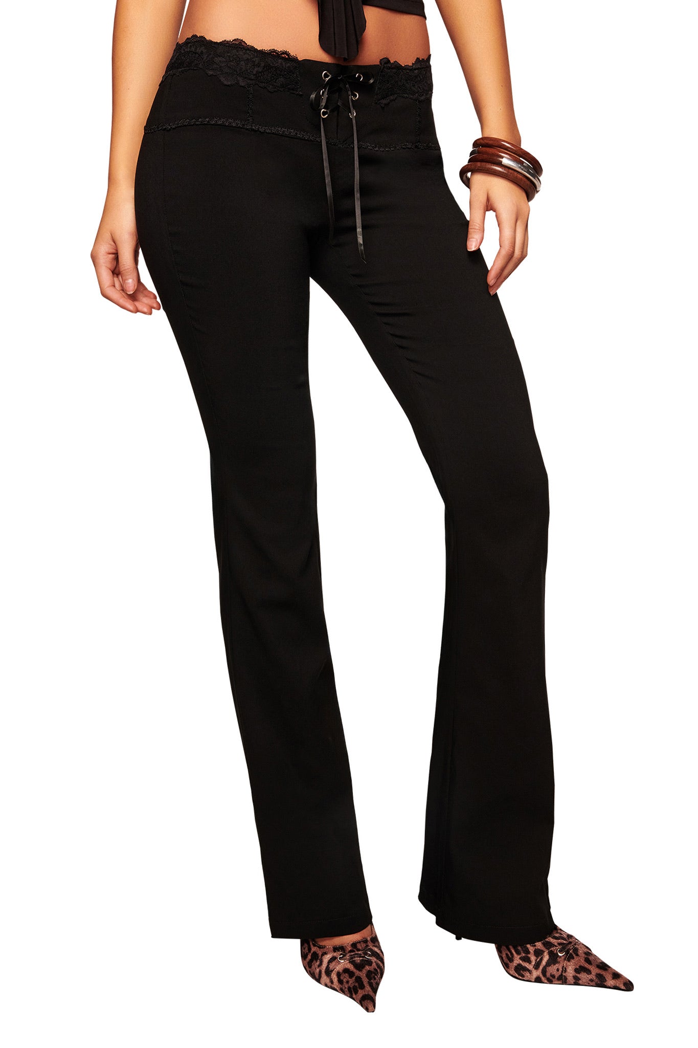 A woman wears I.AM.GIA SERAYA PANT in black with lace trim and a drawstring waist, styled with leopard print pointed-toe shoes and brown bangles. The photo is cropped from the shoulders down.