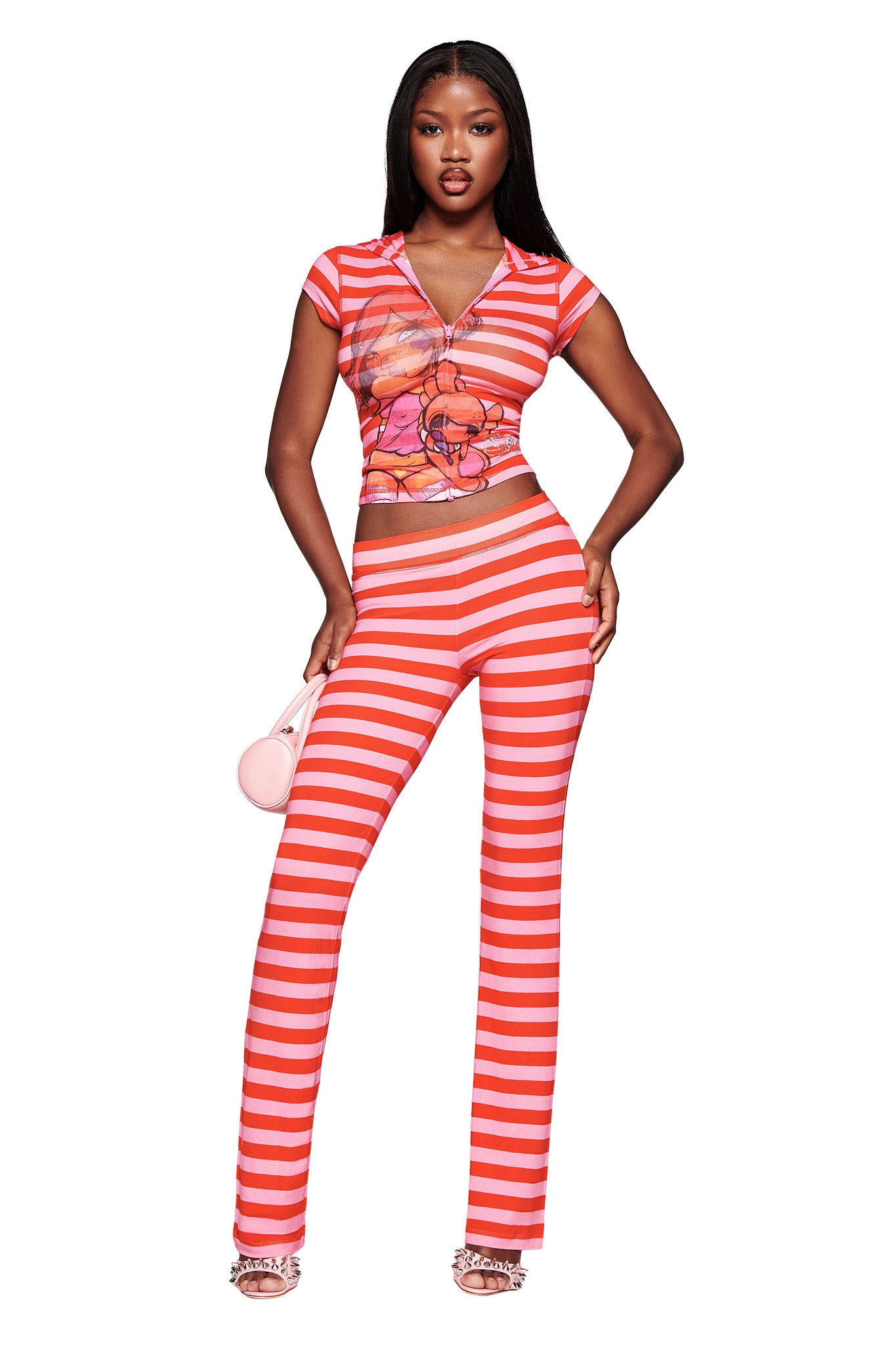 A woman poses in the I.AM.GIA FAFI BLARE TRACKPANT - PINK STRIPE, styled with a matching graphic crop top, pink purse, and heeled sandals. The look features bold red and white stripes and zipper details.