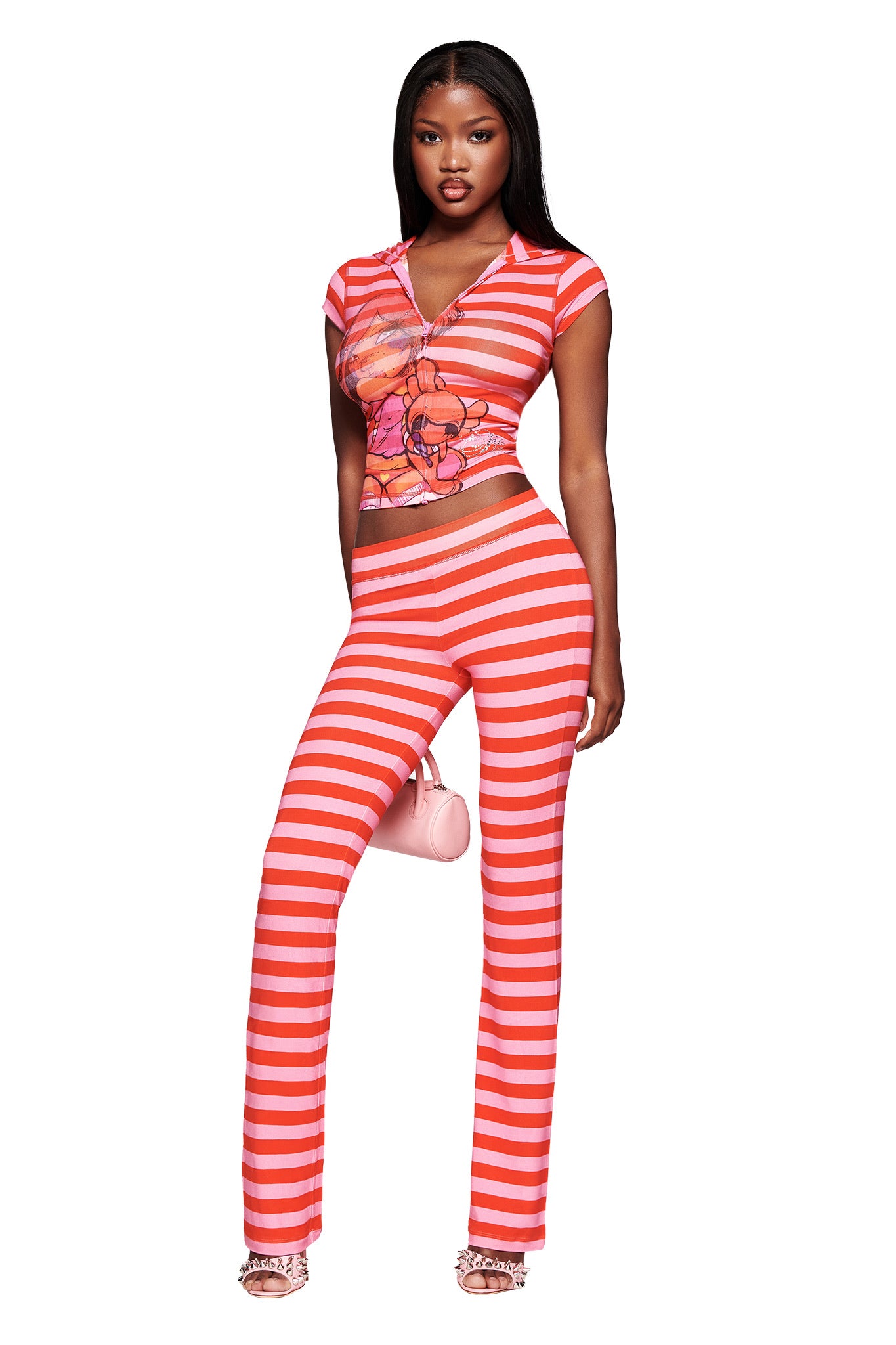 A woman stands confidently in I.AM.GIA’s FAFI BLARE TRACKPANT - PINK STRIPE, styled with a fitted pink graphic bamboo jersey top, strappy heels, and a small pink bag against a white background.