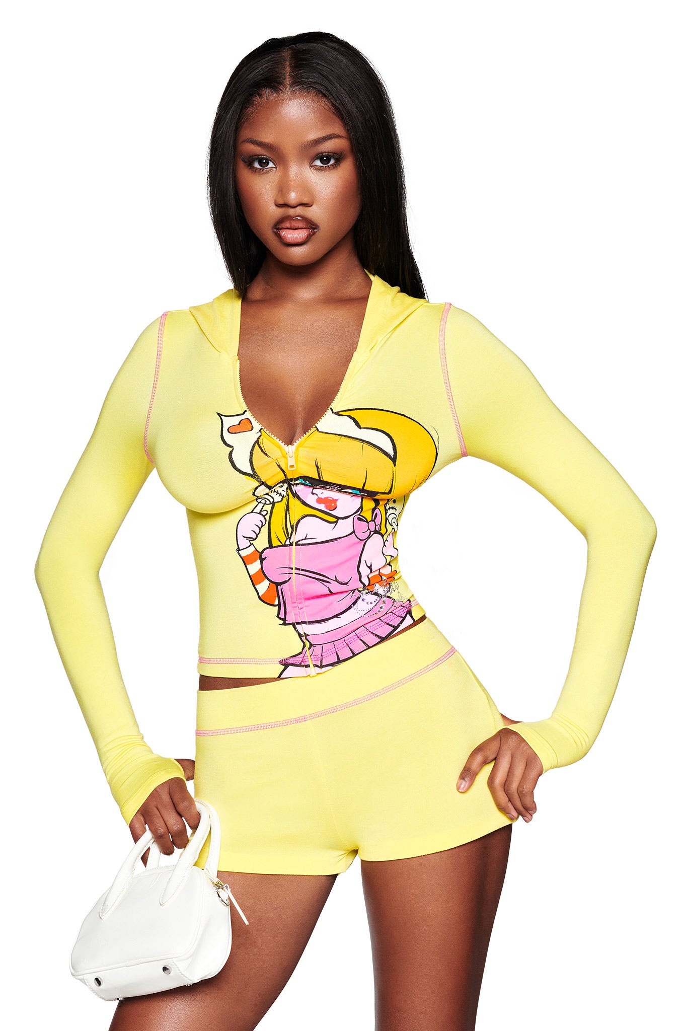 A woman wears the I.AM.GIA FAFI BLARE HOODIE in yellow, featuring pink trim and a Fafinette-inspired graphic, standing with a small white purse against a white background.