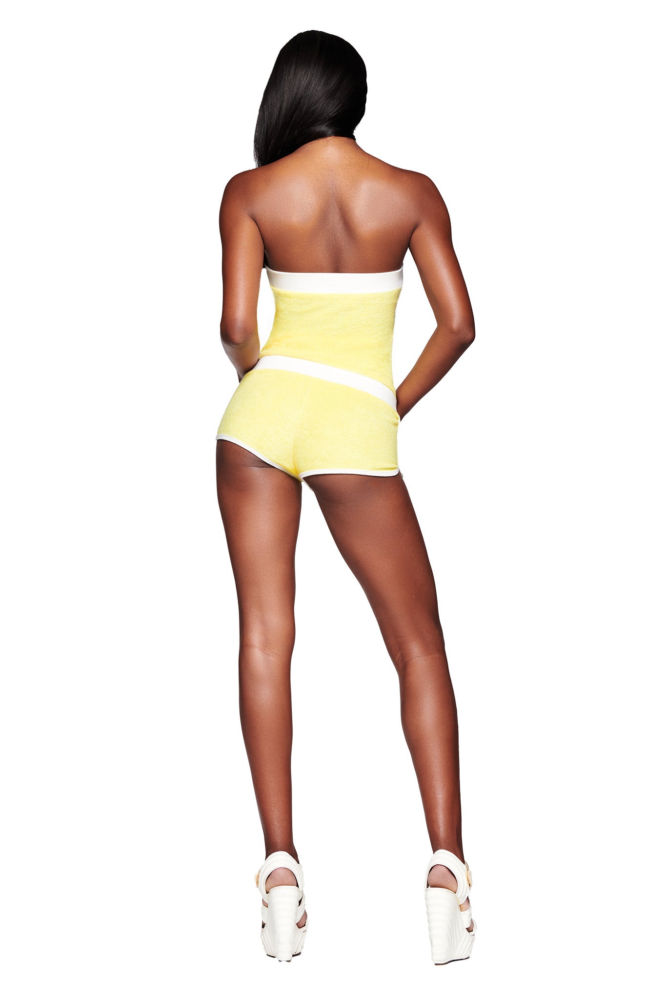 A woman faces away against a white background, wearing the I.AM.GIA GOLDIE ROMPER in yellow with white platform heels, her long hair cascading down her back.