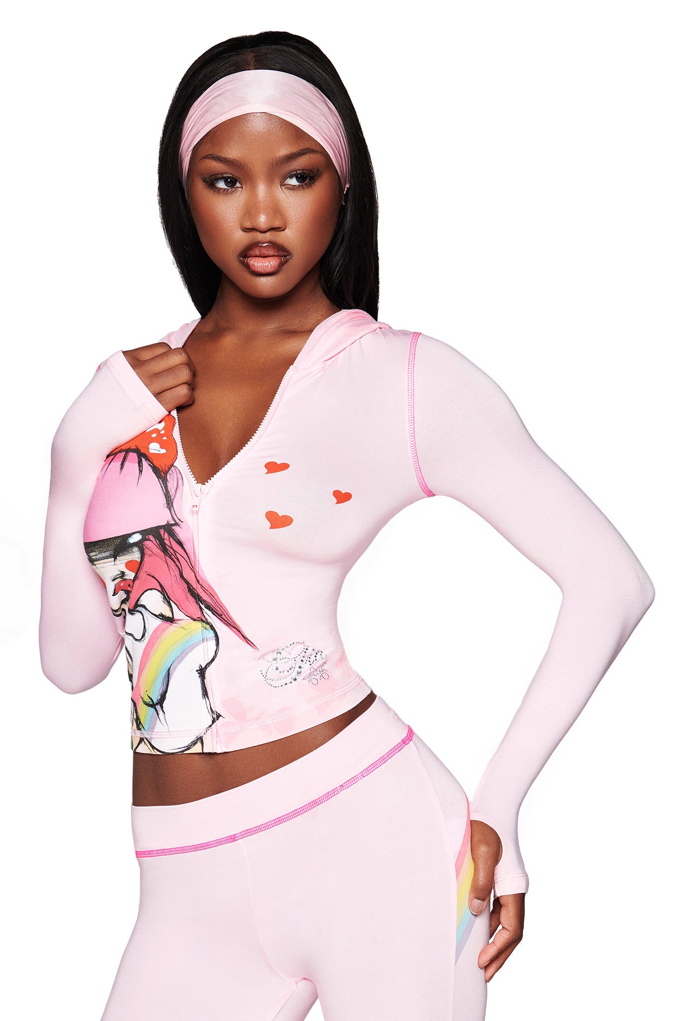 A woman wears the I.AM.GIA FAFI BLARE HOODIE in baby pink, featuring graphic art and heart motifs from the FAFI collab, paired with matching pink leggings with a rainbow detail. She poses confidently against a white background.