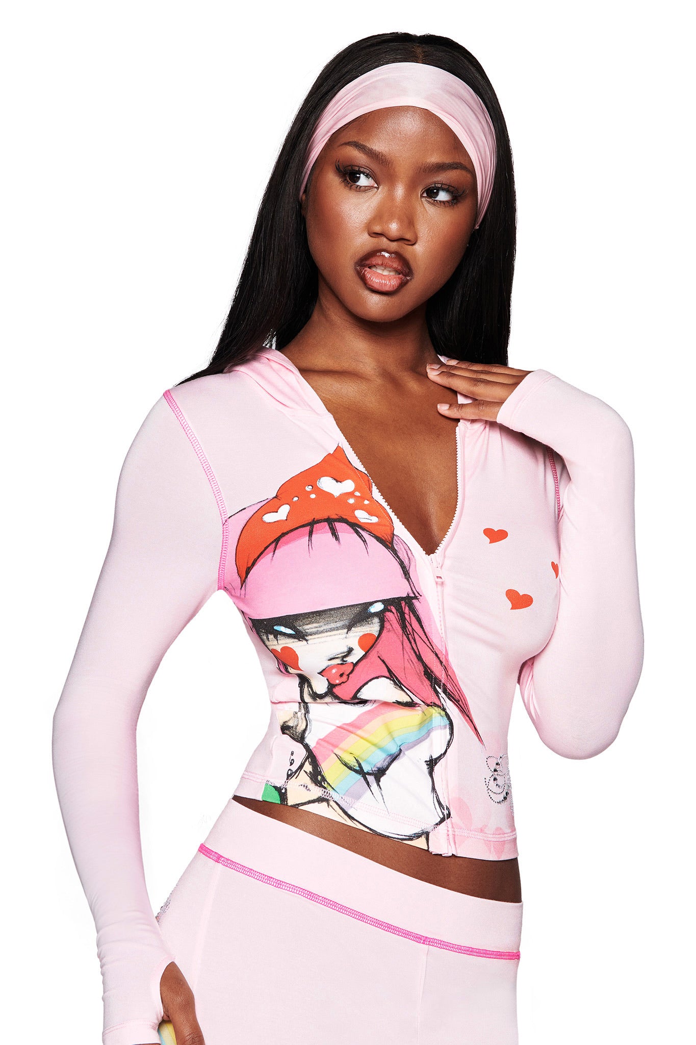 A woman wears the I.AM.GIA FAFI BLARE HOODIE in baby pink, featuring an illustrated girl graphic, red hearts, and pink details. She poses with a hand near her face, highlighting her pink headband and body-con silhouette.