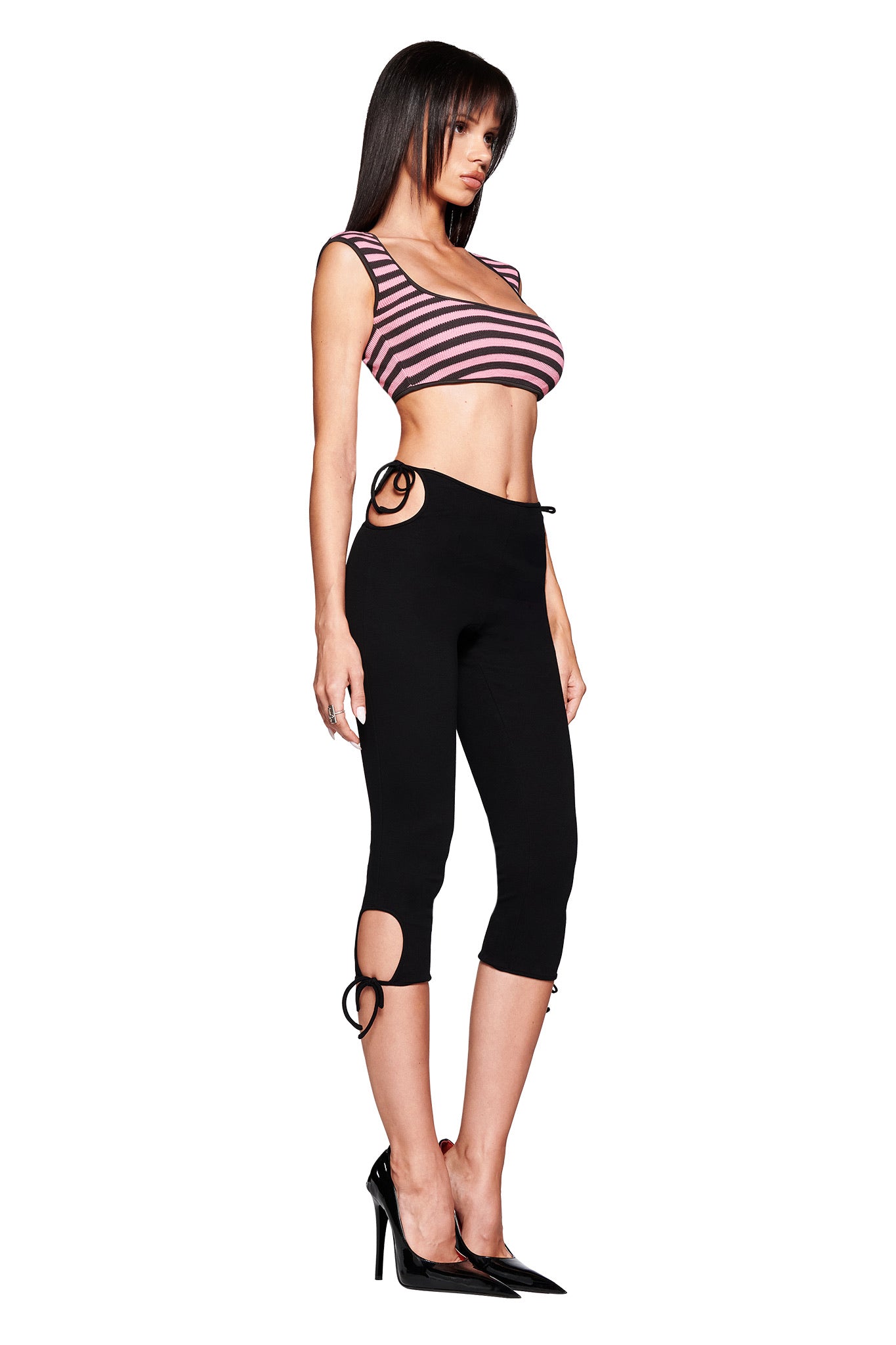 A woman stands in profile against a white background wearing the I.AM.GIA RICA TOP in pink and grey, paired with black capri leggings featuring side cutouts and ring details, and black high heels.