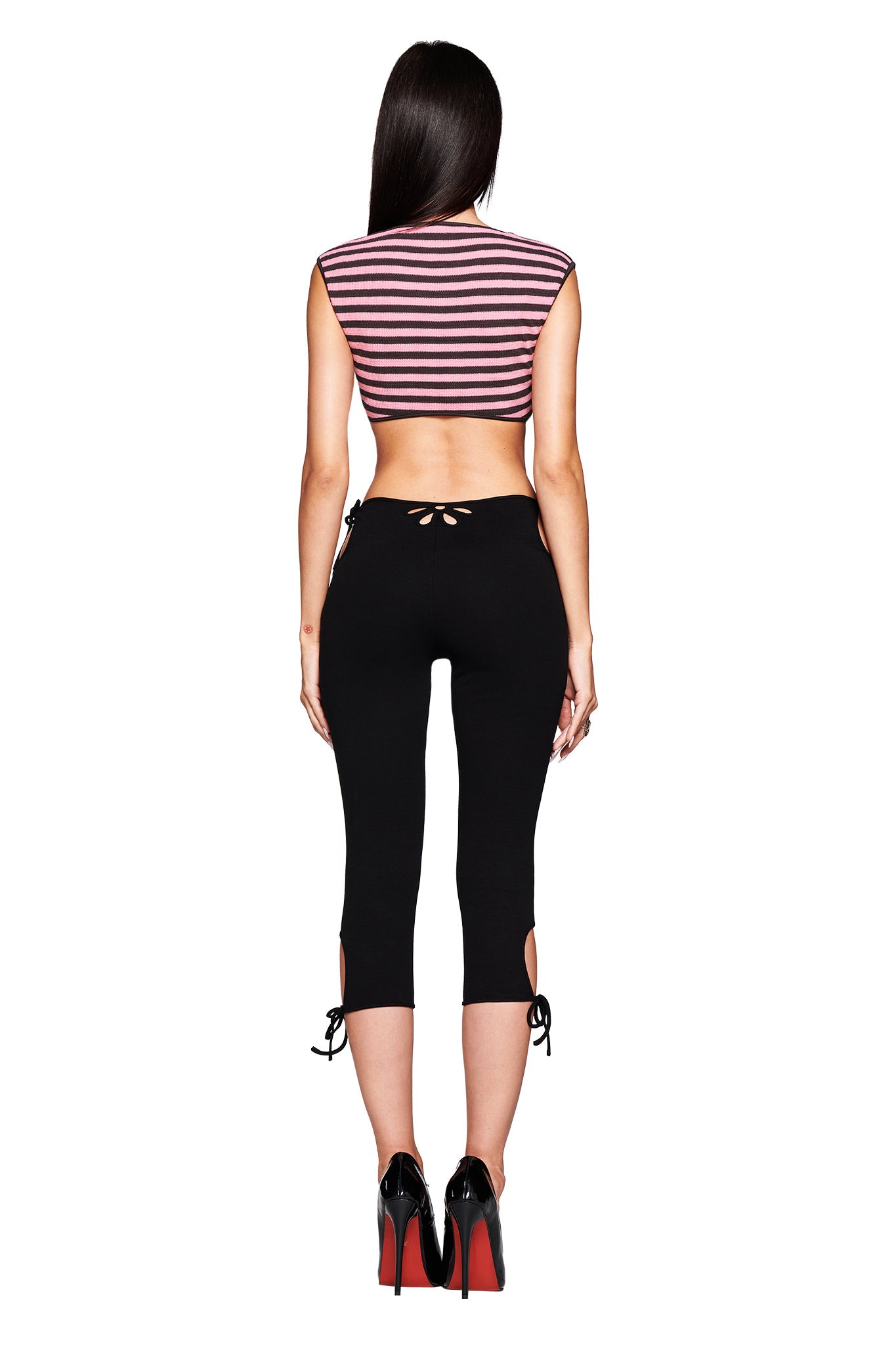 A woman faces away from the camera, wearing the I.AM.GIA RICA TOP in pink and grey, styled with black body-con capri pants featuring cut-outs and bows, plus black high heels with red soles.