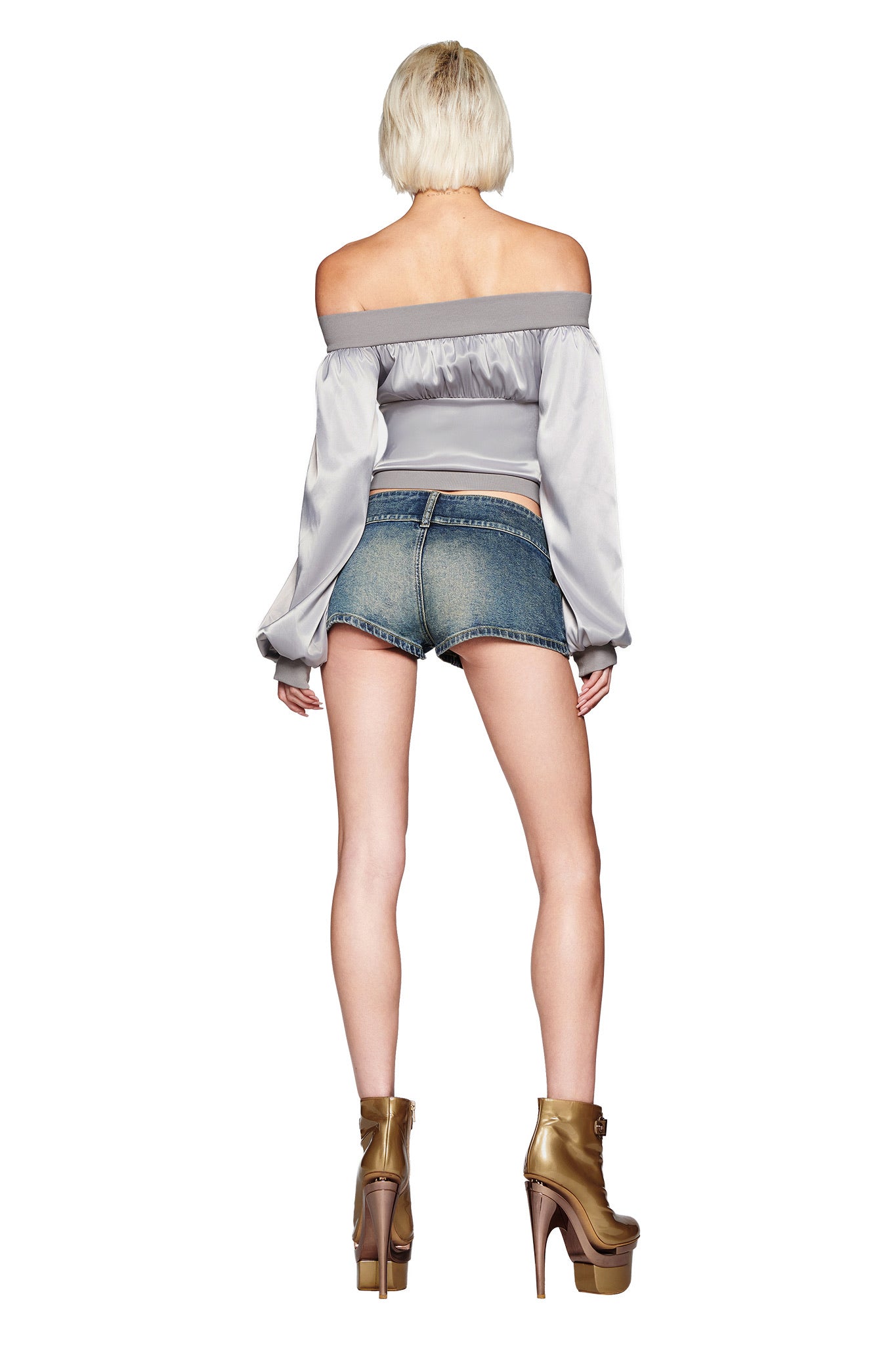 A person with short blonde hair stands with their back to the camera, wearing the I.AM.GIA JORDANA TOP in silver, denim shorts, and gold platform heels against a white background.