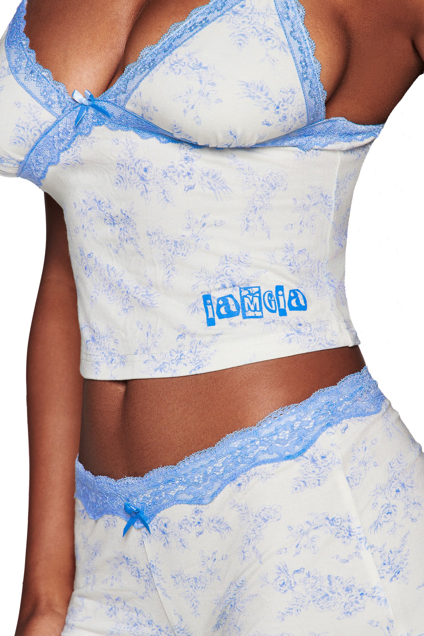 A person wears the KENDRA TANK - BLUE TOILE by I.AM.GIA, a light-colored toile floral camisole with blue lace trim, "iamgia" in blue, and adjustable straps. The image highlights the torso and matching shorts.