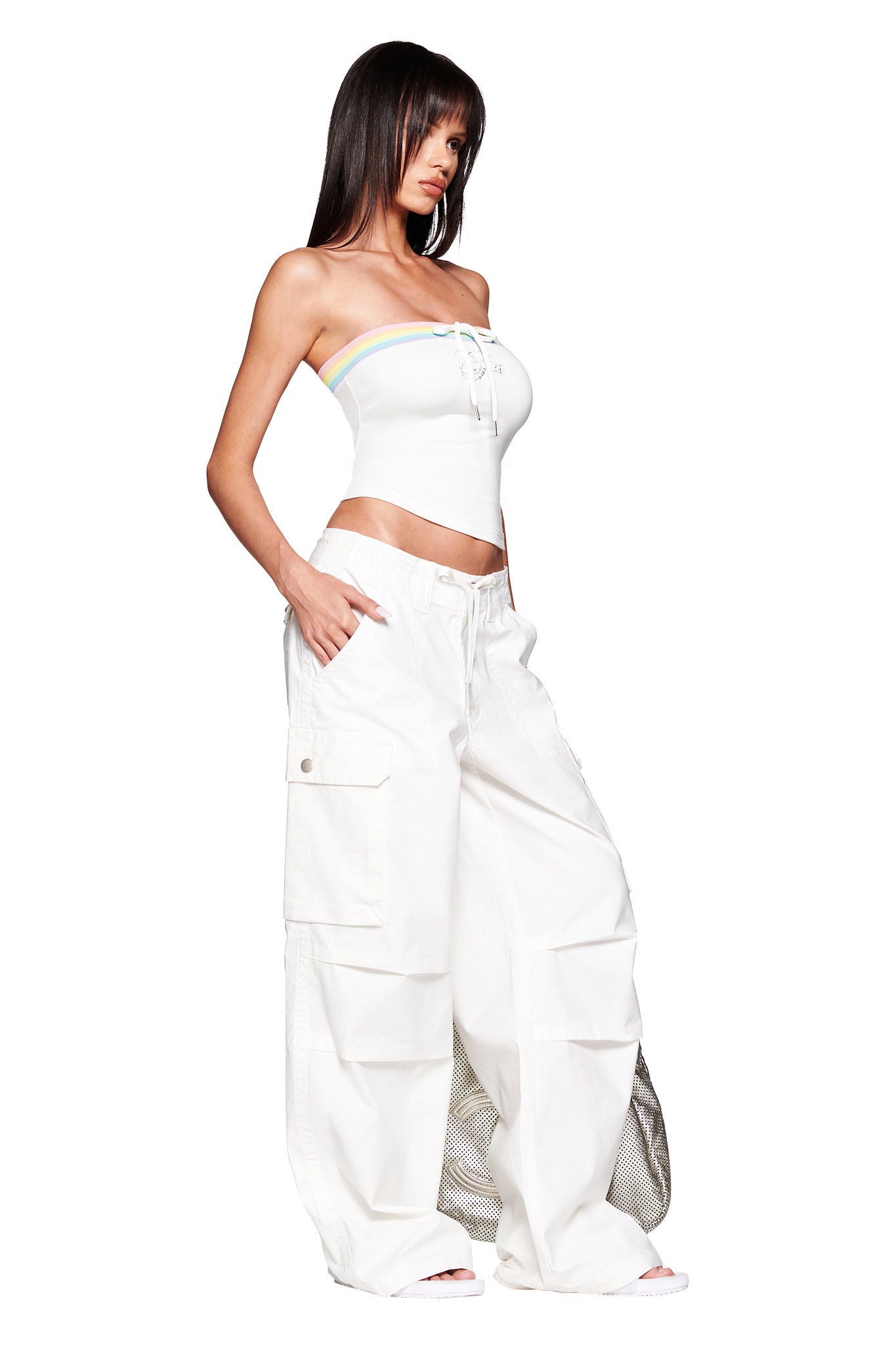 A woman wears the I.AM.GIA NIJI BOOB TUBE - WHITE, featuring a pastel stripe and lace-up front, paired with loose white cargo pants and sandals, posing against a plain white background.
