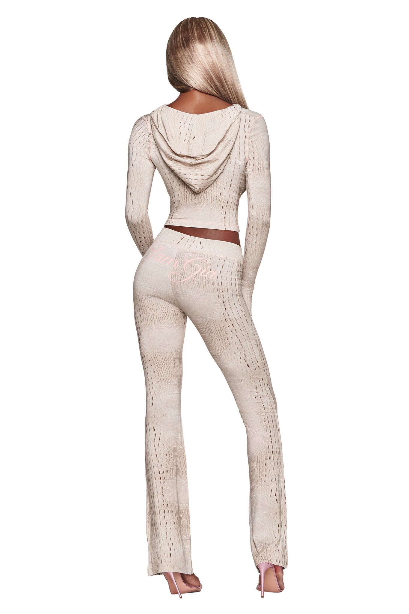 A person with long blonde hair is seen from the back, wearing a beige knit hooded top and matching I.AM.GIA BLARE TRACKPANT - HOT CROC flared pants, paired with high heels against a white background.