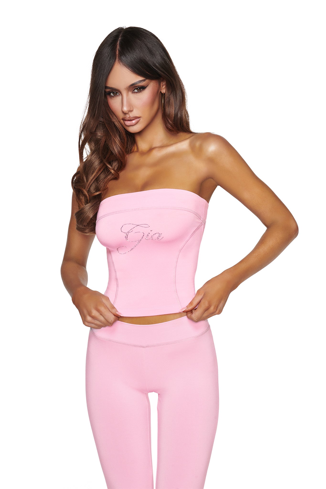 A woman with long brown hair wears the I.AM.GIA BLARE DIAMONTE BOOB TUBE in baby pink—a strapless crop top adorned with a sparkling “Gia” rhinestone logo—paired with matching pants against a white background. She holds the bottom edge of the tube top.