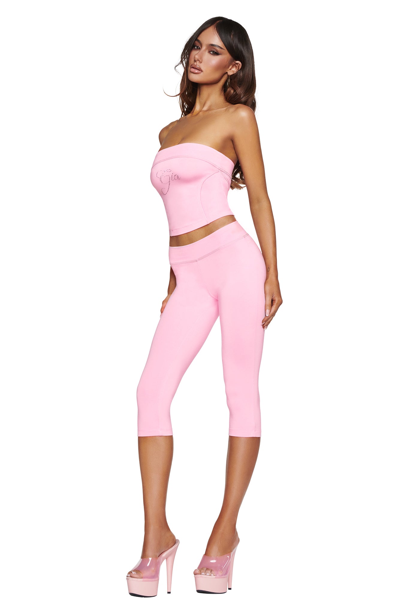 A woman poses against a white background wearing the I.AM.GIA BLARE LOW RISE DIAMONTE CAPRI in baby pink, styled with a strapless pink top and matching pink high-heeled platform shoes.
