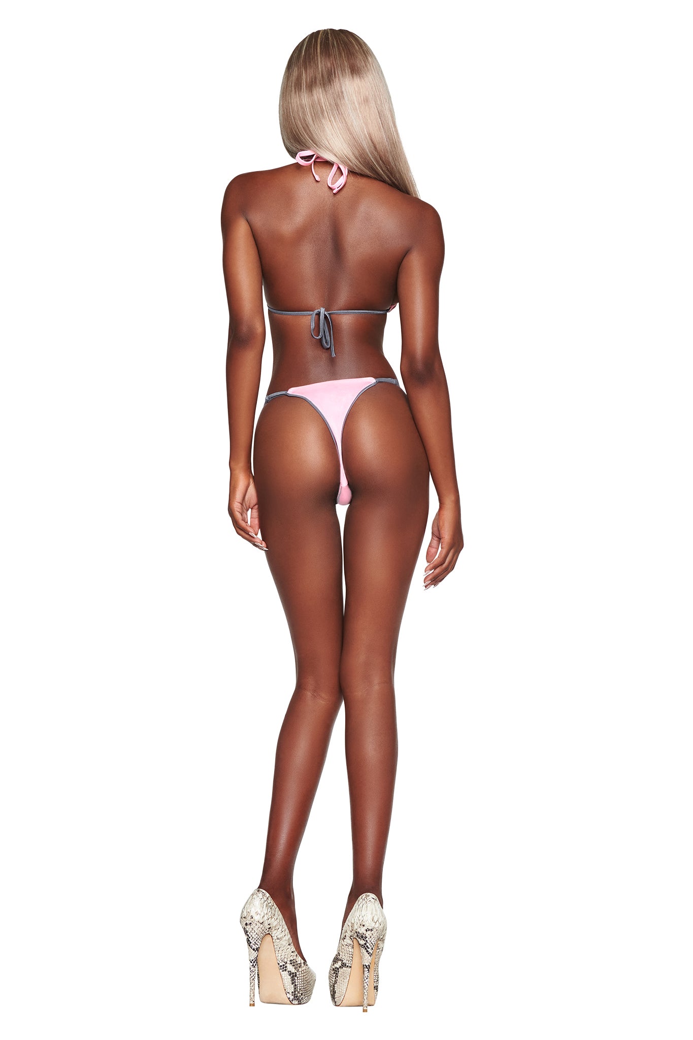 A woman with long blonde hair, wearing the I.AM.GIA CAMERON BIKINI BOTTOM in baby pink and high heels, stands facing away from the camera against a white background.