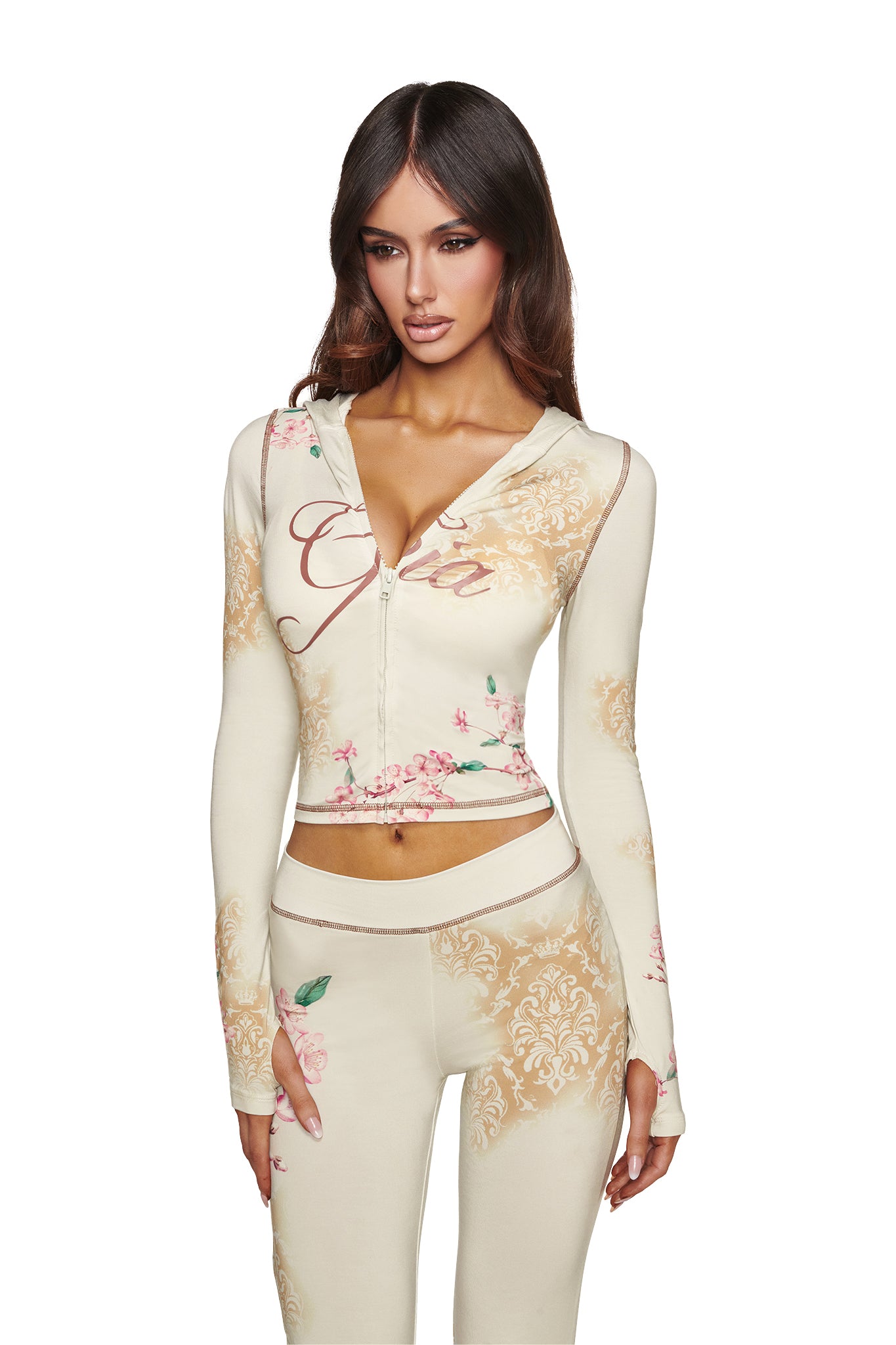 A woman wearing the I.AM.GIA BLARE HOODIE - BLOSSOM—a cream-colored, fitted hoodie with matching pants featuring blossom floral prints and cursive text—stands against a white background.