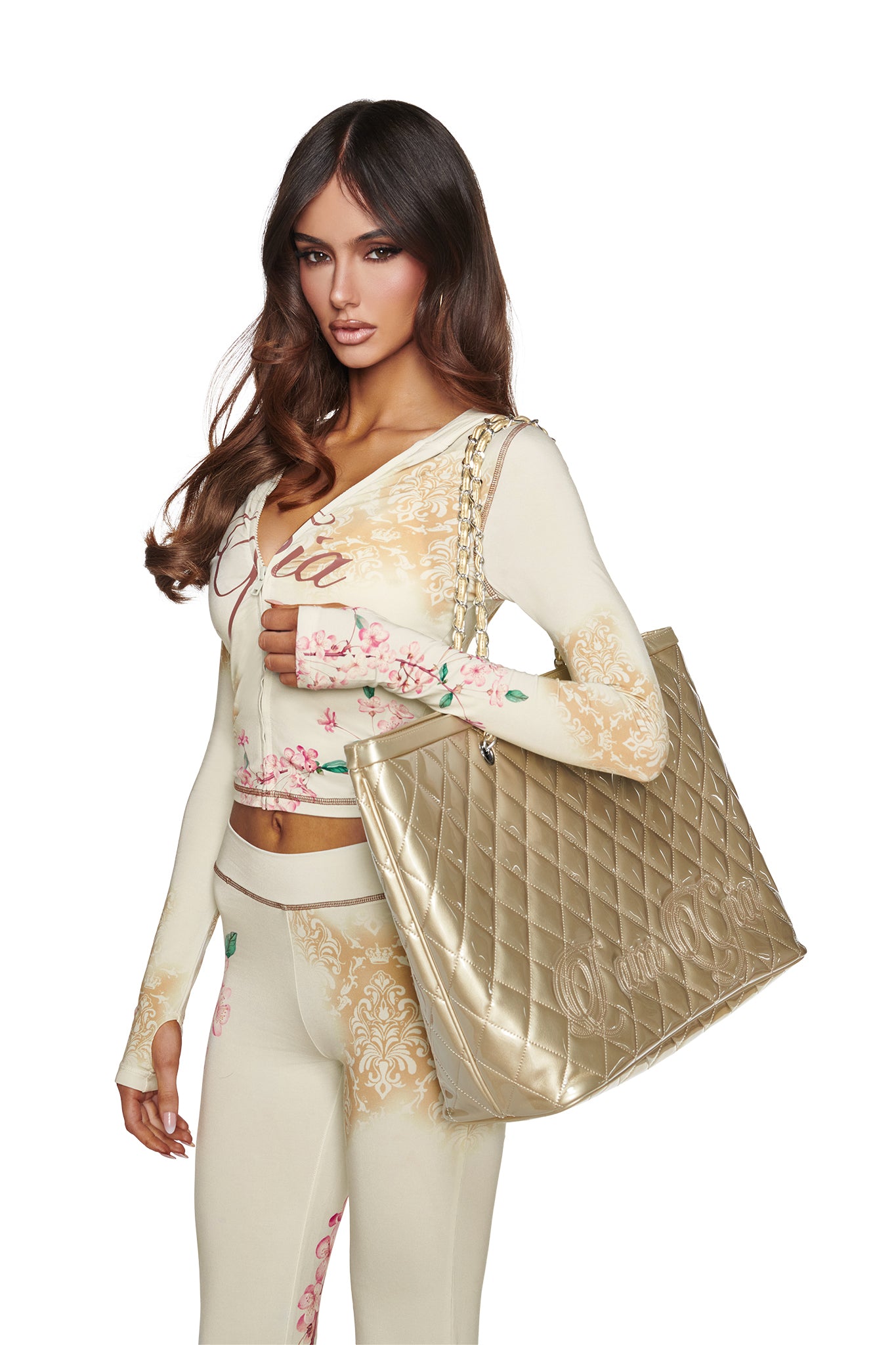 A woman with long brown hair wears a floral-patterned cream outfit and carries the I.AM.GIA DANNI TOTE BAG in gold over her shoulder against a white background.