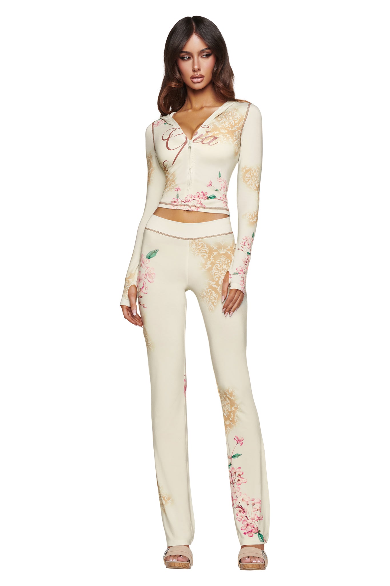 A woman stands against a plain white background wearing the I.AM.GIA BLARE HOODIE in a cream-colored blossom print, paired with matching pants. She has long brown hair and is wearing sandals.