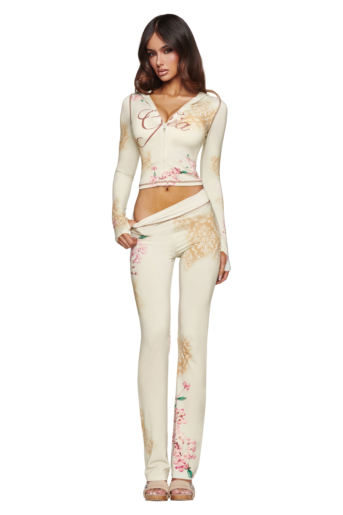 A woman wearing the I.AM.GIA BLARE HOODIE in Blossom, a cream-colored, blossom print two-piece outfit with long sleeves and flared pants, poses against a white background.