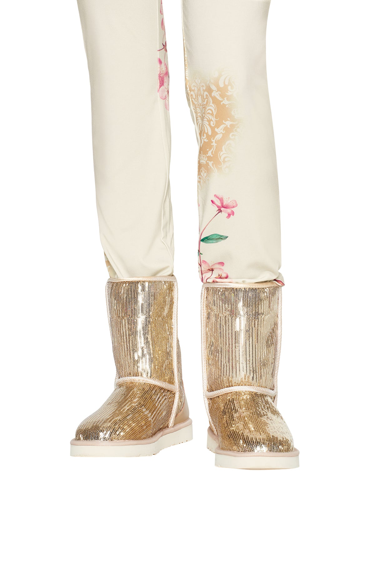 A person wearing cream-colored floral pants and the I.AM.GIA ELVIRA SEQUIN BOOT in gold, featuring shiny sequins and a cozy faux fur lining, stands on a white background.