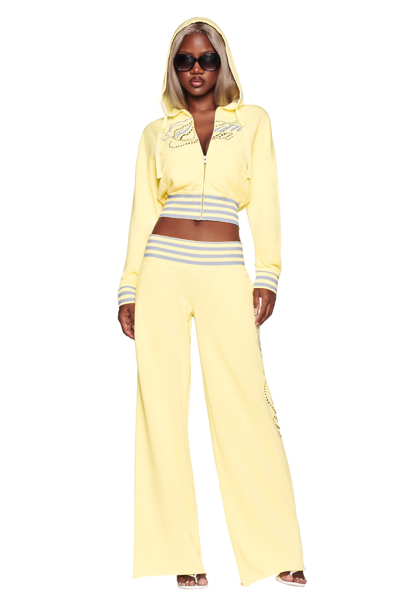 A woman stands against a white background wearing the I.AM.GIA VEDA PANT in yellow—a light yellow, low-rise, wide-leg tracksuit pant embellished with rhinestone detailing—paired with a matching hooded top that shows her midriff, sunglasses, and heeled sandals.