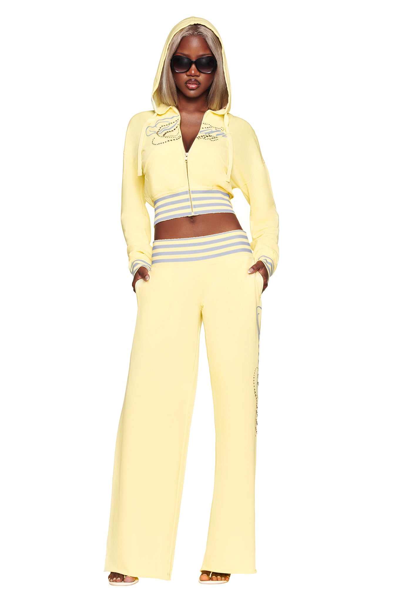 A woman wears the I.AM.GIA VEDA JACKET in yellow—a pale zip-up cropped hoodie with wide-leg pants featuring gray stripes and an oversized GIA script rhinestone detail. She completes the look with sunglasses and heels, posing with her hands in her pockets against a white background.