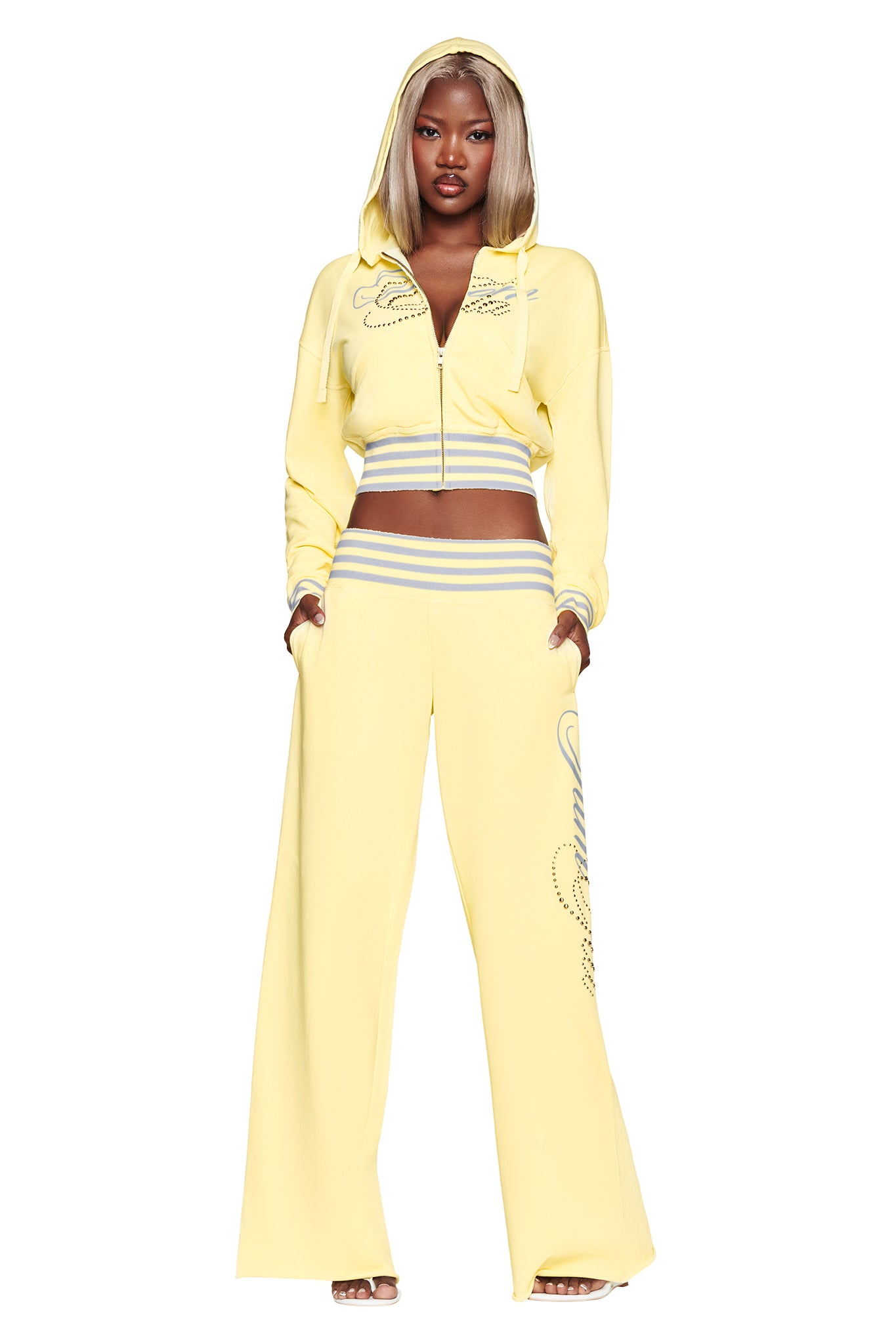 A woman stands confidently against a white background wearing the I.AM.GIA VEDA PANT in yellow—a wide-leg silhouette with a gray and white waistband—paired with a light yellow hooded crop jacket.