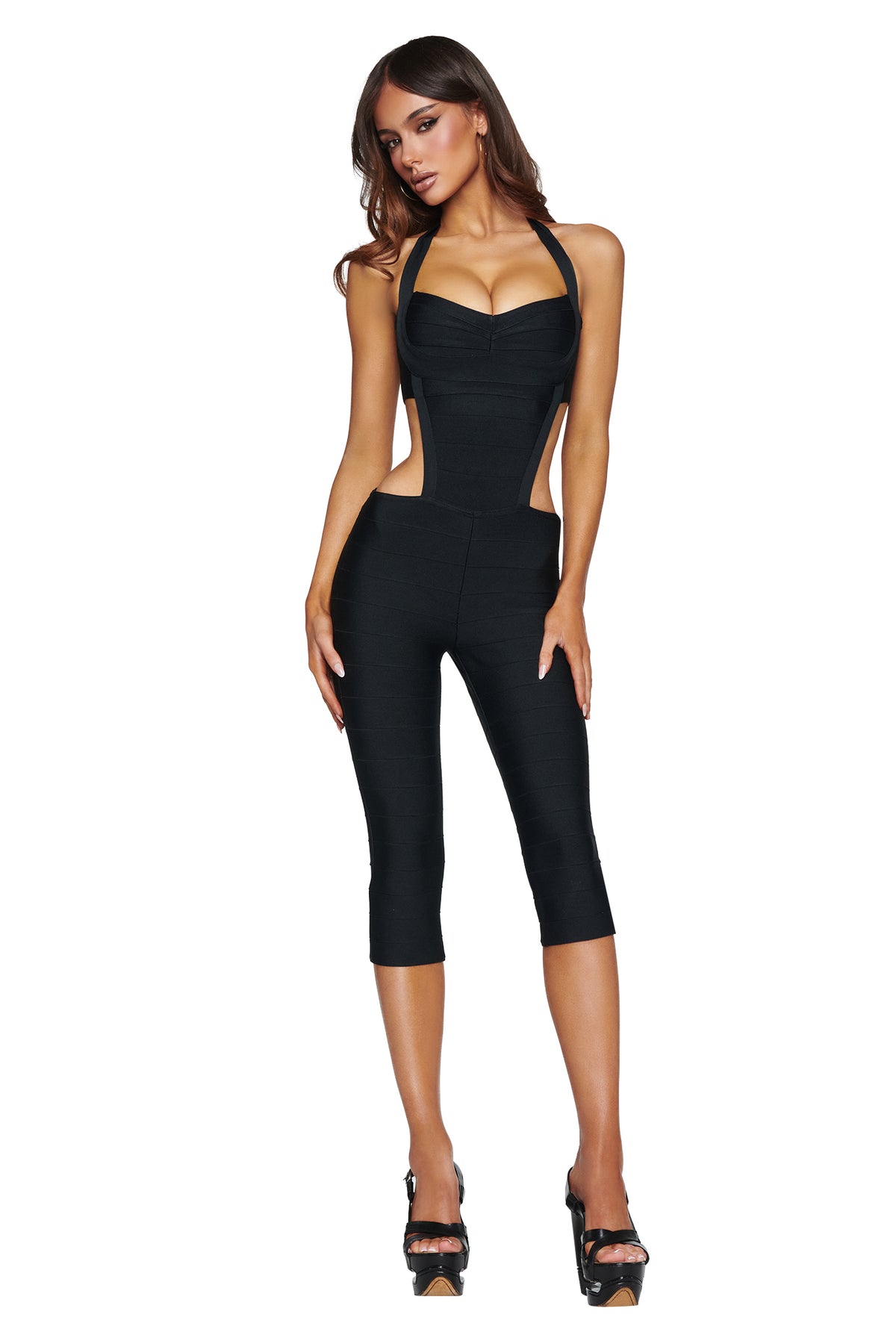 A woman models the I.AM.GIA EVA Bandage Capri Jumpsuit in black, featuring a halter neck, cutout sides, and cropped legs, styled with black high heels against a white background.