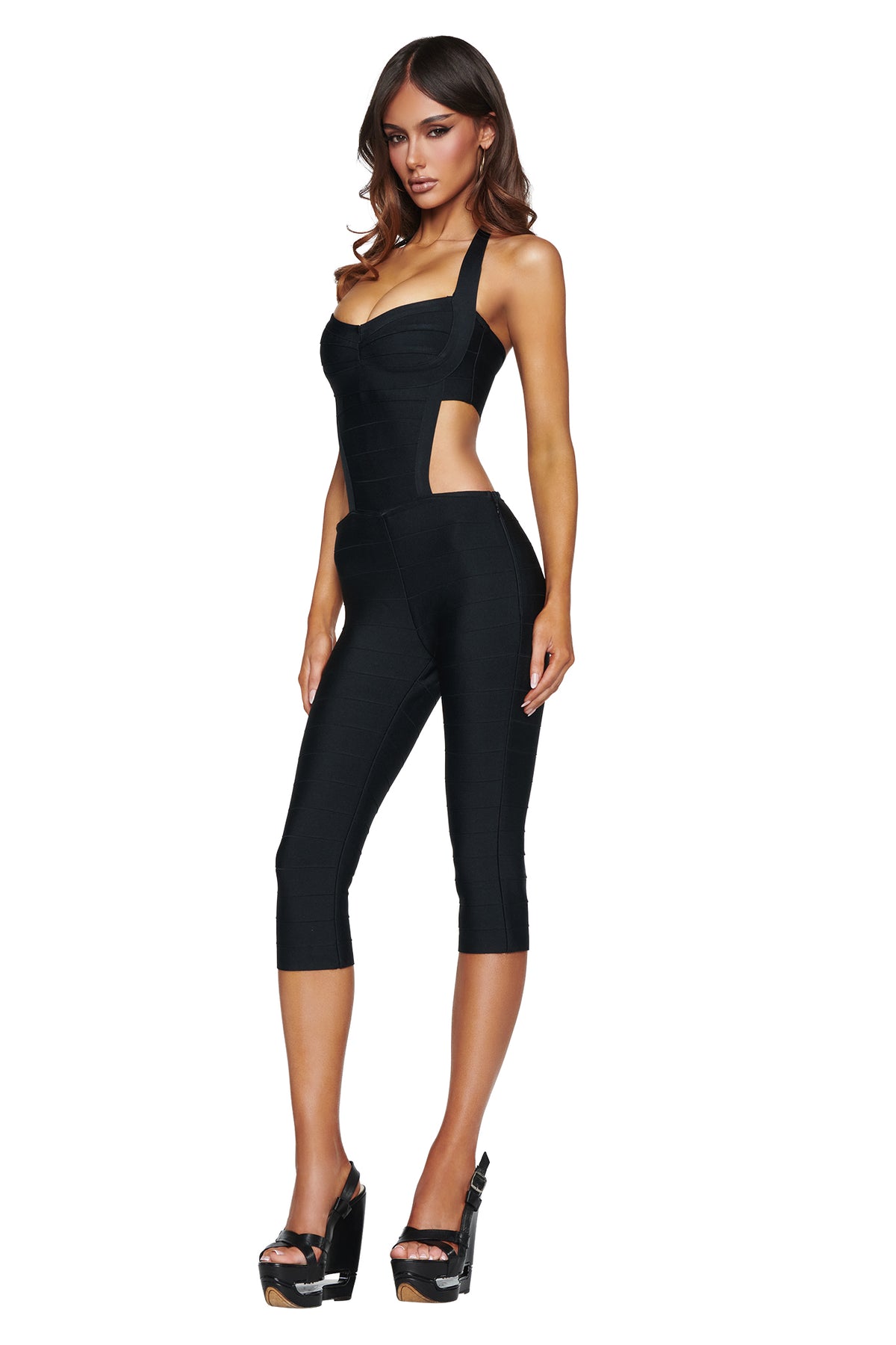 A woman wearing the I.AM.GIA EVA BANDAGE CAPRI JUMPSUIT in black, featuring side cutouts and capri-length legs, stands in high platform heels against a white background.