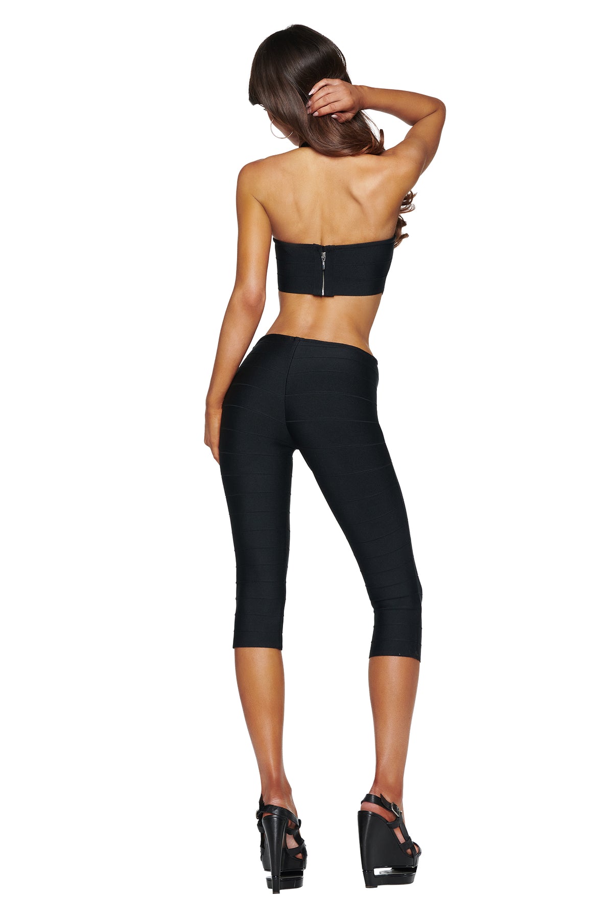 A woman models the I.AM.GIA EVA Bandage Capri Jumpsuit in black, standing with her back to the camera, hand in her hair, and wearing black platform heels against a white background.