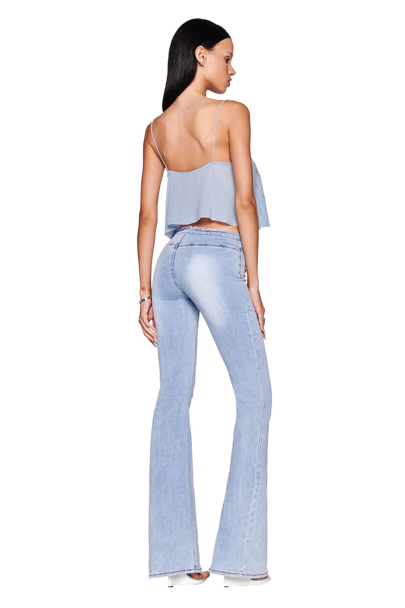 A woman in an I.AM.GIA baby blue MINA TOP and flared jeans stands with her back to the camera. Her long dark hair is adorned with butterfly diamante artwork, and she's wearing transparent high-heeled shoes against a white background.