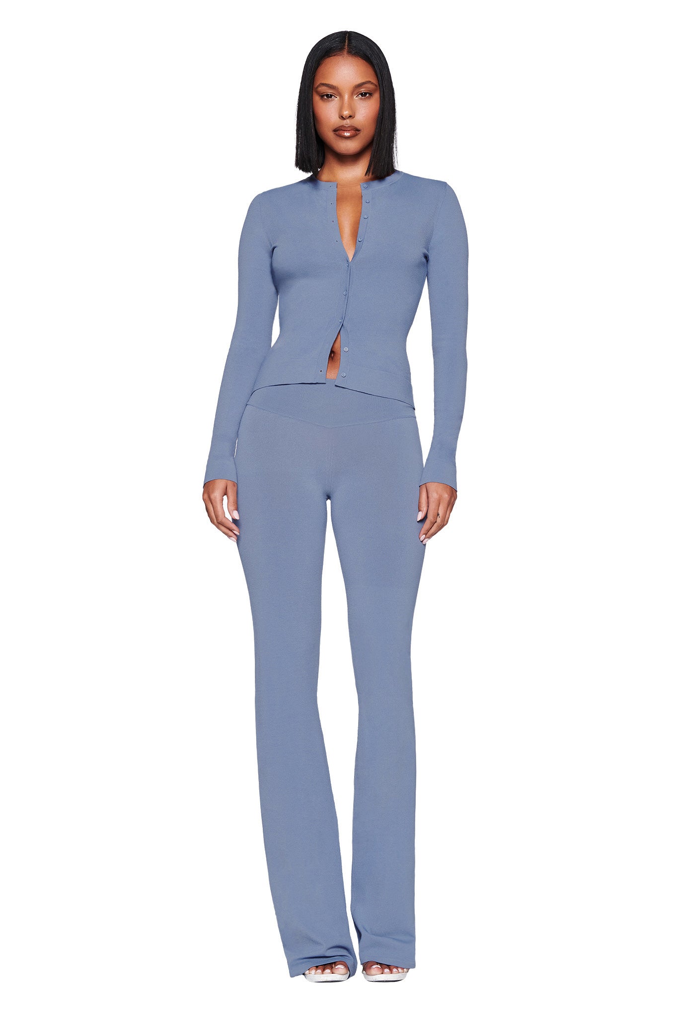 A woman, her bob hairstyle complementing her look, stands against a white background wearing a fitted long-sleeve blue top with buttons and matching I.AM.GIA Audrina Pant in steel blue knit. She finishes the outfit with white open-toed shoes.