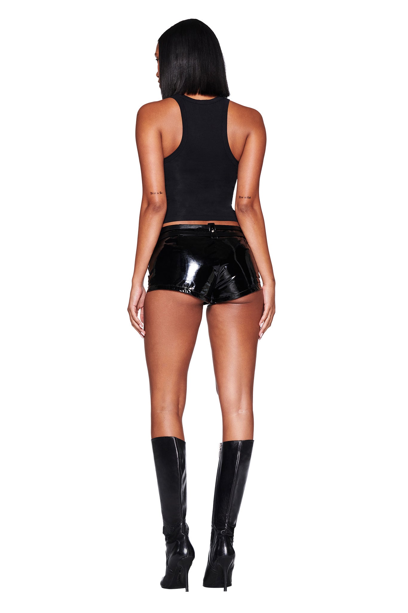 A person with long dark hair faces away from the camera, wearing a black sleeveless top and I.AM.GIA's VEGA HOT SHORT in black, paired with knee-high black boots against a white backdrop.