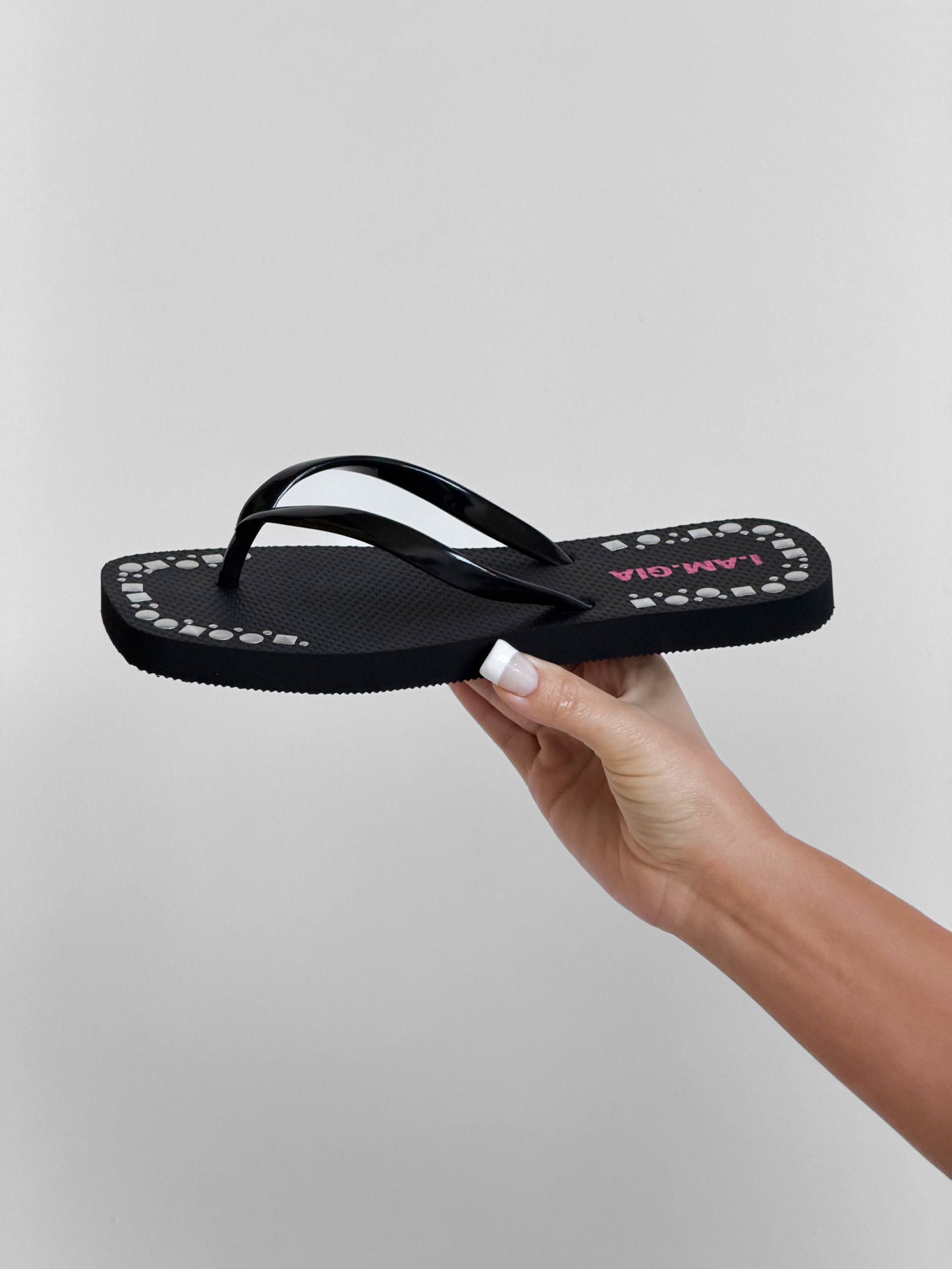 A manicured hand holds the INKA STUDDED FLIP FLOP - BLACK by I.AM.GIA, a black sandal with a white and pink patterned strap, stud details, and an extra-soft foam footbed, set against a plain light background.
