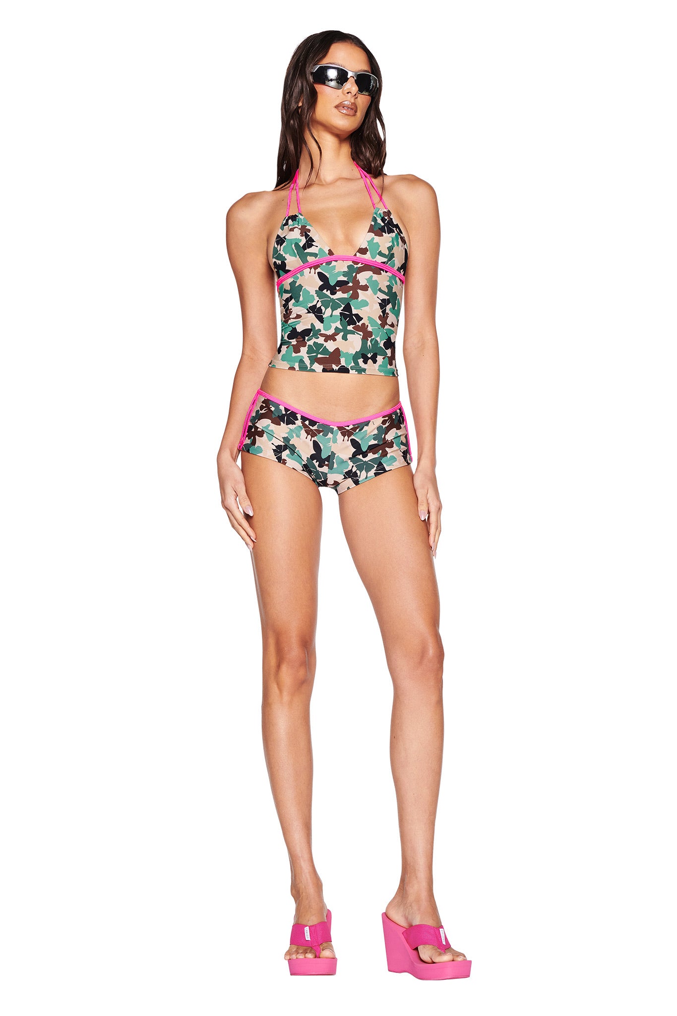 Against a plain white backdrop, Joanne Short confidently flaunts the I.AM.GIA Flutter Camo tankini with pink accents, matching shorts, and pink platform sandals. Bold sunglasses complete her look.