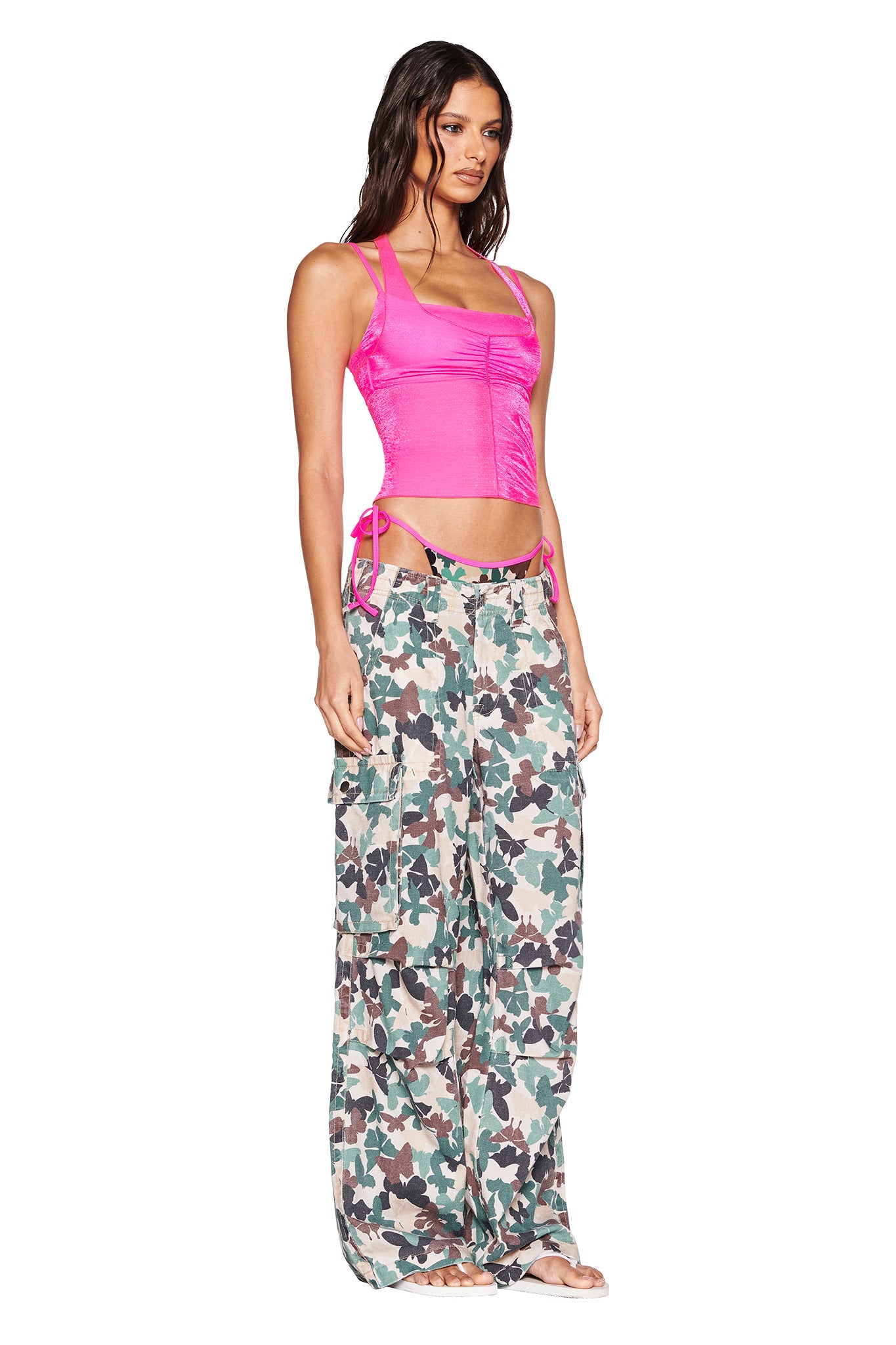 A woman in the I.AM.GIA NERINA TOP - NEON PINK and loose camouflage pants stands against a white background. She wears open-toed shoes, with long wavy hair cascading down her shoulders.