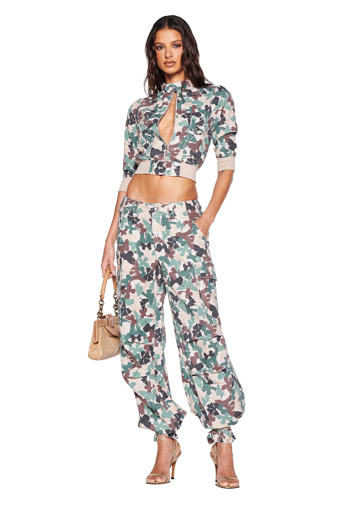 A woman models the I.AM.GIA EFFIE JACKET in flutter camo, paired with matching pants. She completes her look with a keyhole neckline top, beige handbag, and gold heels. Her hands rest in her pockets, highlighting her long, wavy hair with flair.